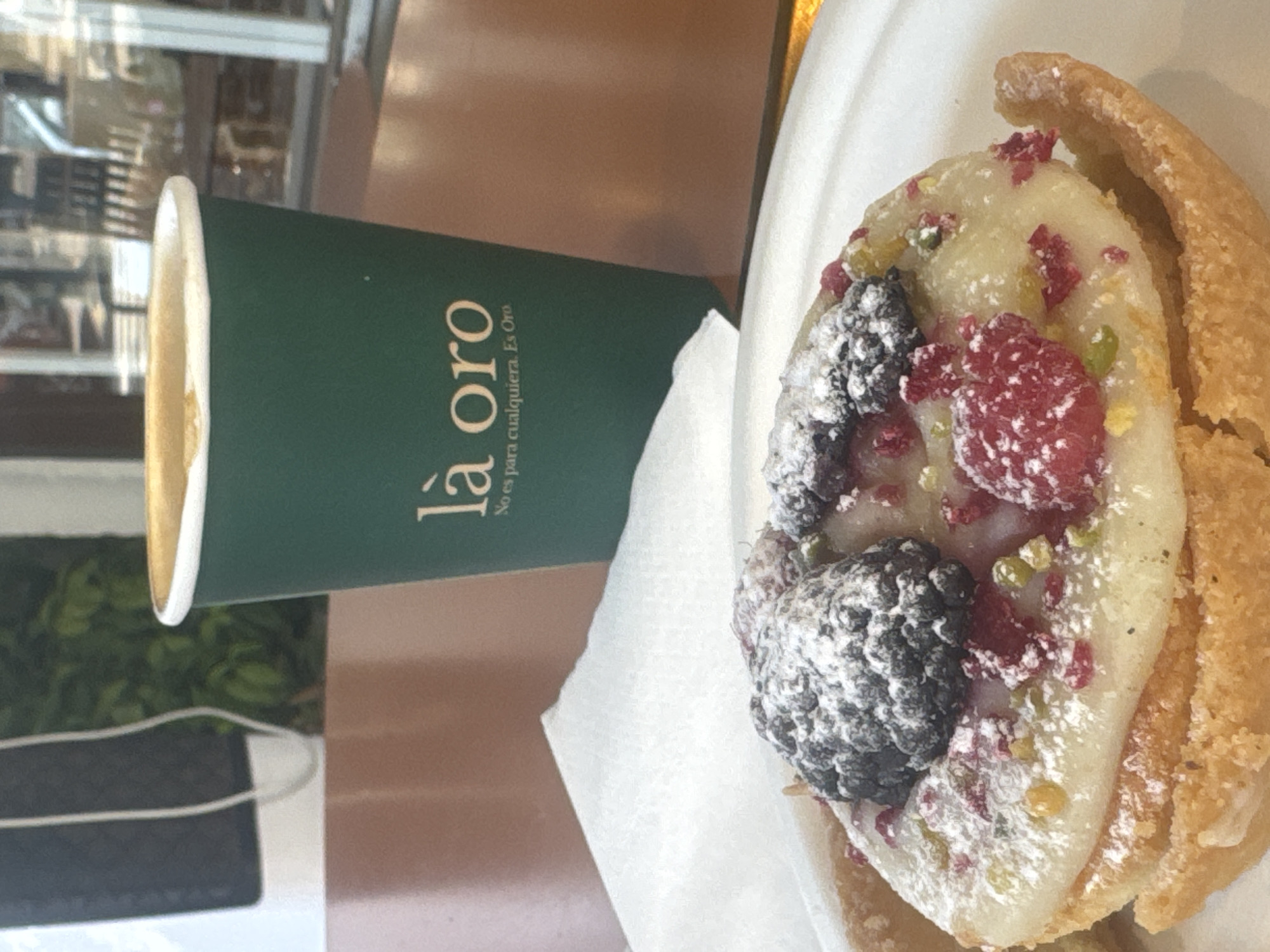 A close-up shot focuses on a pastry topped with white icing, a blackberry, raspberries, and chopped pistachios, all dusted with powdered sugar. The pastry sits on a white plate, partially obscuring a white napkin and a dark brown table. In the background, a green paper cup with gold lettering that reads "là oro No es para cualquiera. Es Oro." is prominent. Beyond the cup, a blurred window shows reflections of shops and greenery, suggesting an urban or commercial setting. The image is likely taken in Ramat Gan, Israel, given the context.