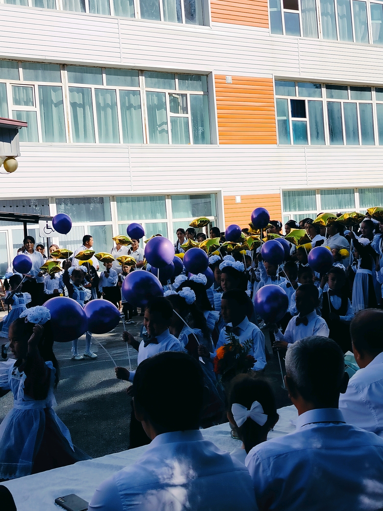 In the city of Kölkent, Kazakhstan, on a bright day, a gathering of formally dressed children, many in school uniforms, are holding purple balloons and star-shaped gold balloons. The children are positioned in front of a modern, multi-story building with white horizontal siding and large windows. The building's exterior also features an orange-colored section. Some adults are present among the children, appearing to be supervising or participating in the event. The ground is paved, suggesting an outdoor school or public space. The scene appears to be a celebration or a formal school event, possibly the beginning of the school year.