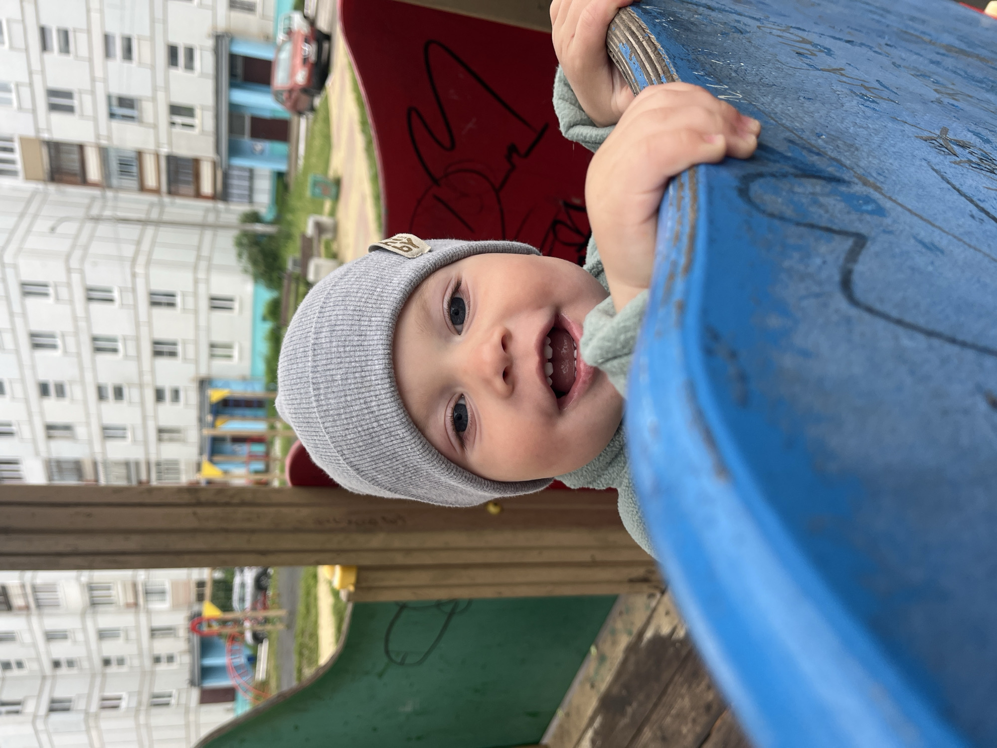 The image is oriented on its side, featuring a young child peeking from the left side of the frame. The child, likely a toddler, looks directly at the camera with wide blue eyes and an open-mouthed, joyful expression, revealing some lower teeth. They wear a light grey knit beanie with a small, indistinct patch on the front, and a light green, ribbed long-sleeved top. Their small hands grasp the worn edge of a bright blue playground slide that runs diagonally across the right side of the image.
The blue slide shows signs of extensive use with scratches, faded paint, and some unidentifiable black graffiti-like markings. Behind the child and the blue slide, a portion of a red playground structure with similar black graffiti is visible. The background, softly out of focus, reveals an urban residential area in Chaikovsky, Russia. Multiple-story, light-colored apartment buildings with numerous windows dominate the distant view. Patches of green grass, a paved area, and a red car are also discernible among the background elements. The scene captures a playful moment on a playground, likely during a daytime outing with soft, overcast lighting. There is no legible text visible on any of the surfaces.