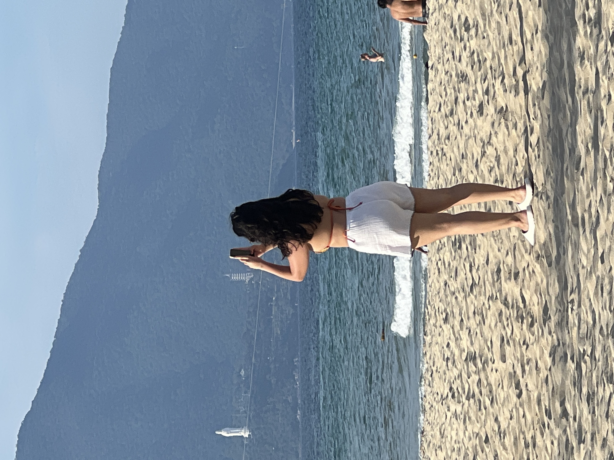 A woman stands on a sandy beach, facing away from the camera, holding a smartphone to take a picture. She is wearing a bikini top and white shorts. The ocean is calm with small waves rolling onto the shore. In the distance, a large mountain range dominates the horizon, and a white statue and a pagoda are visible on its slopes. The sky is clear and blue. Several other people are visible in the water and on the beach in the background. The scene is likely located at My Khe Beach in Da Nang, Vietnam, given the presence of the Marble Mountains and the Lady Buddha statue.