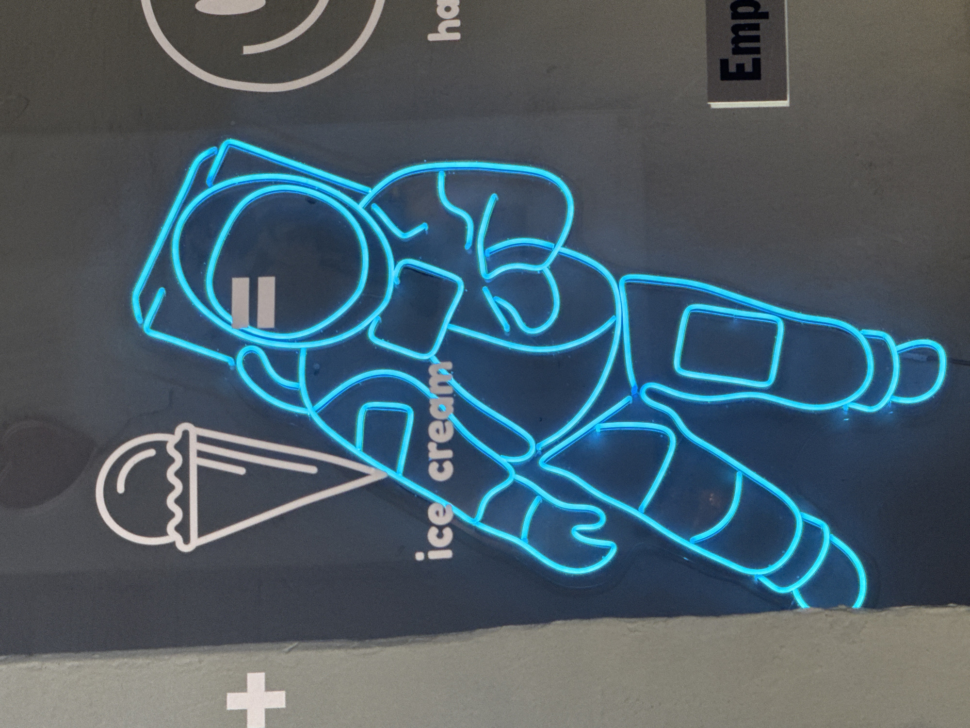 A close-up shot of a gray wall with neon signs, possibly in the city of Querétaro, Mexico, based on the presence of Spanish text. The central element is a bright blue neon sign depicting an astronaut in a spacesuit, angled diagonally across the frame. Adjacent to the astronaut, to the left, is a white outline of an ice cream cone. The words "ice cream" are written in a white sans-serif font, positioned vertically alongside the ice cream cone. Above the astronaut is a partial view of a white smiley face outline. To the right of the smiley face is the barely visible word "ha". Further right and almost out of frame is the word "Emp", likely the start of a word such as "employee." Below the ice cream cone is a white plus sign. The bottom edge of the image shows a lighter, possibly gray, surface. The overall impression is a playful, modern design commonly found in commercial spaces.
