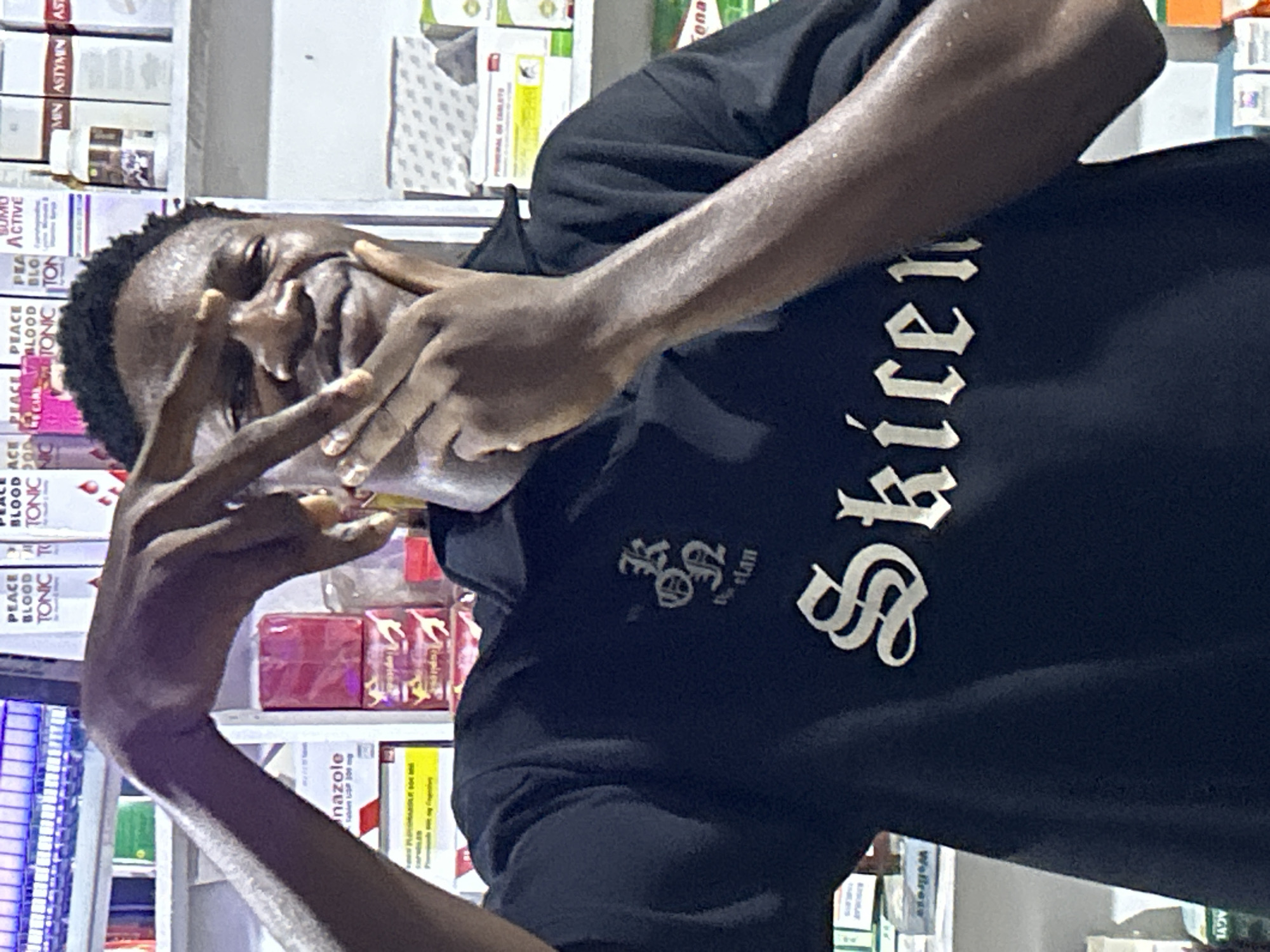 A young, dark-skinned man is posed in a narrow, indoor space resembling a pharmacy or dispensary. He is wearing a black hoodie with the word "Skicem" in a Gothic-style font. The location appears to be Ihiagwa, Nigeria. The background consists of shelves filled with medications and health products. The man's face is partially obscured by his hands, which are forming a "peace" or "victory" sign. He is smiling, although his eyes are somewhat obscured. The lighting is bright and uneven.