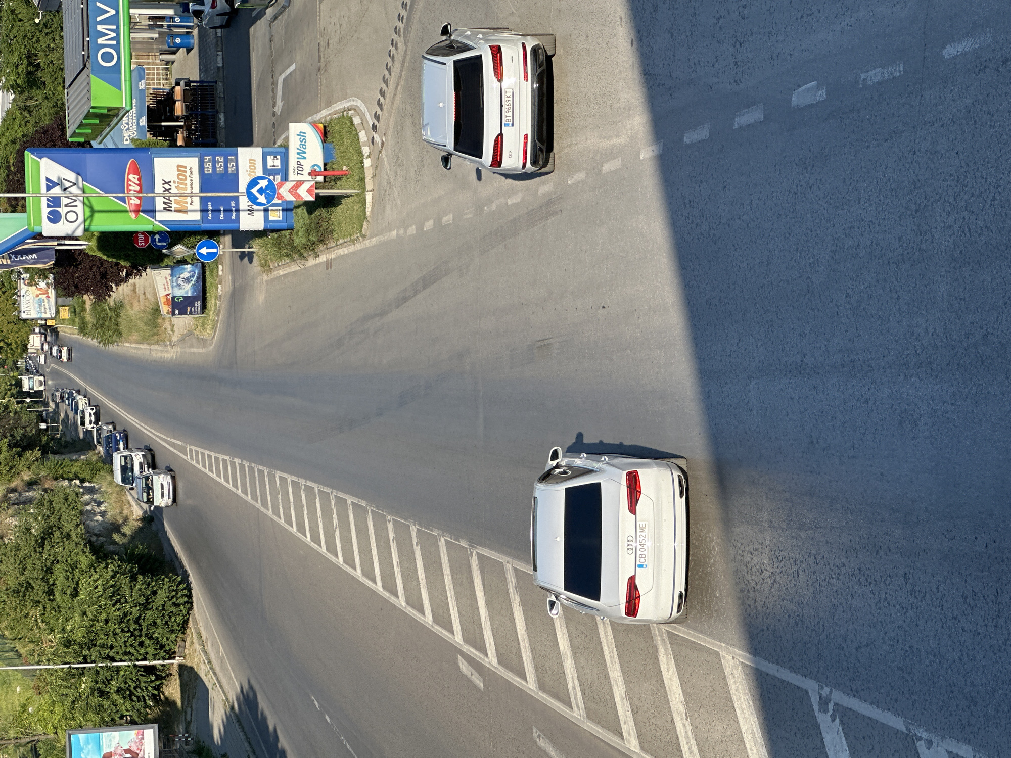 This is a top-down, vertically oriented photograph of a street scene in Veliko Tarnovo, Bulgaria, during daylight. The scene depicts a line of cars, predominantly white SUVs, waiting at a traffic light or possibly queuing for a car wash at an OMV gas station on the right side of the road. The gas station has prominent blue and green signage, with "OMV" and "MAXX Motion" visible. A "Top Wash" sign is also present. The road itself is asphalt with white dashed lane markings.
In the foreground, a white Audi SUV with the license plate "CB 0452 ME" is stopped, casting a shadow across the road. Further down the road, another white Audi SUV, this one with the license plate "BT 9669 KT", is also visible. The surrounding environment includes lush green trees and bushes lining the left side of the road. A billboard advertising "BIANCO" and "XXAM" is partially visible in the lower left corner. The overall impression is a snapshot of everyday traffic and commerce on a sunny day in a Bulgarian city. No people are clearly discernible in the image. The lighting suggests it's likely late afternoon or early morning due to the long shadows.