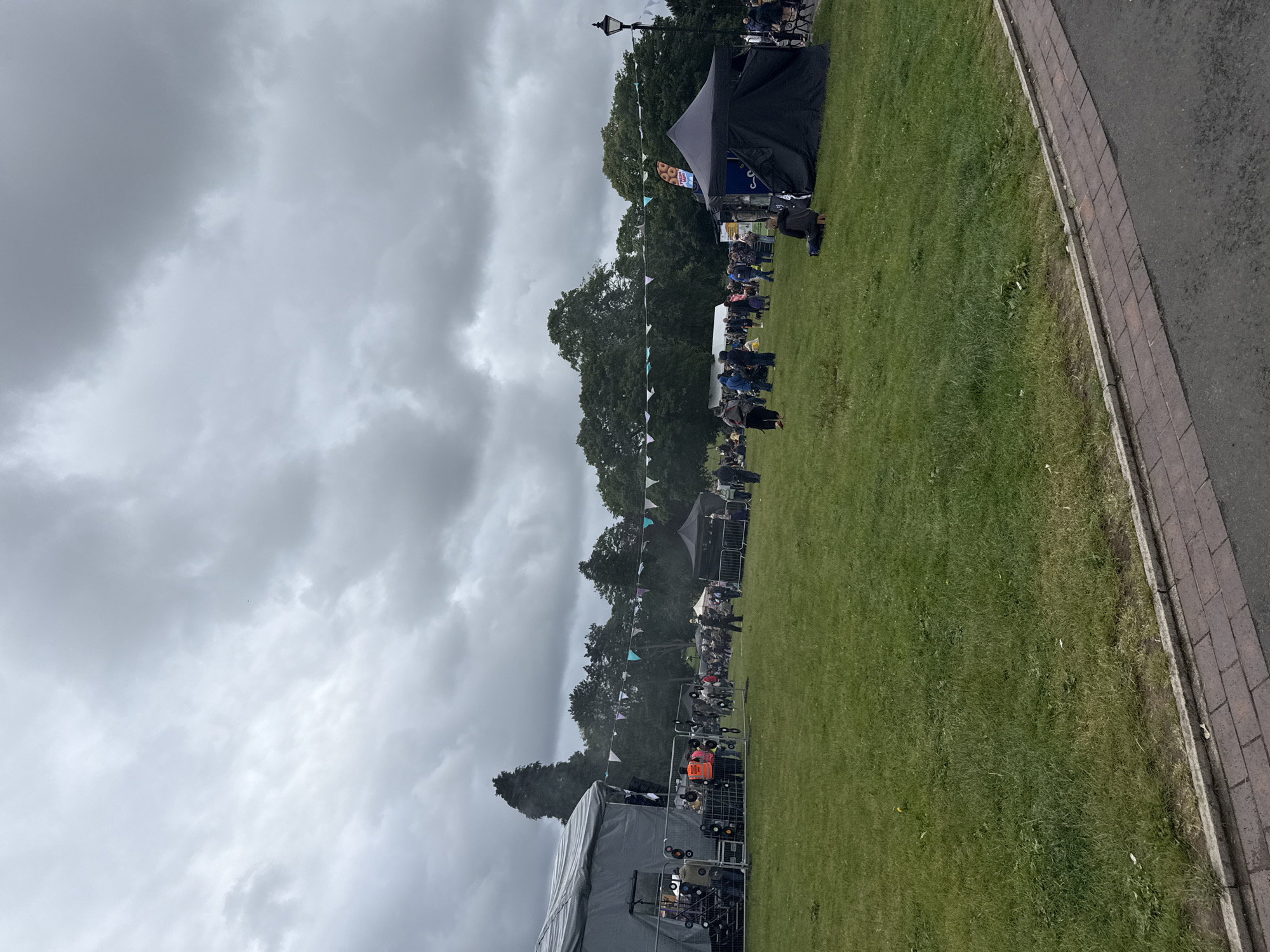 This image captures an outdoor public gathering or festival in a park-like setting, under an heavily overcast sky. The location is an unknown city, in an unknown country. The ground consists of extensive green grass, with a paved pathway composed of asphalt and brick pavers running along the immediate foreground.
In the mid-left, a large, grey, temporary stage structure is erected, enclosed by metal barriers. Several individuals are present around the stage area, including some wearing orange high-visibility vests, suggesting event staff or volunteers. Vinyl records are decoratively attached to the metal fencing around the stage.
Across the midground, numerous people are scattered across the grass. They are engaged in various activities, including standing, walking, and sitting. Many are casually dressed, some wearing jackets or hoodies. Towards the mid-right, a prominent black canopy tent with a peaked roof stands on the grass; a sign partially visible above it advertises "FRESH DONUTS." Further back and to the right, a white tent and a black ornate lamppost are visible.
The background is dominated by a dense treeline of mature green trees, with light-colored triangular bunting strung between them, adding a festive element. The sky above is entirely covered by thick, grey clouds, indicating overcast weather and diffuse light. There are no clear indications of direct sunlight.