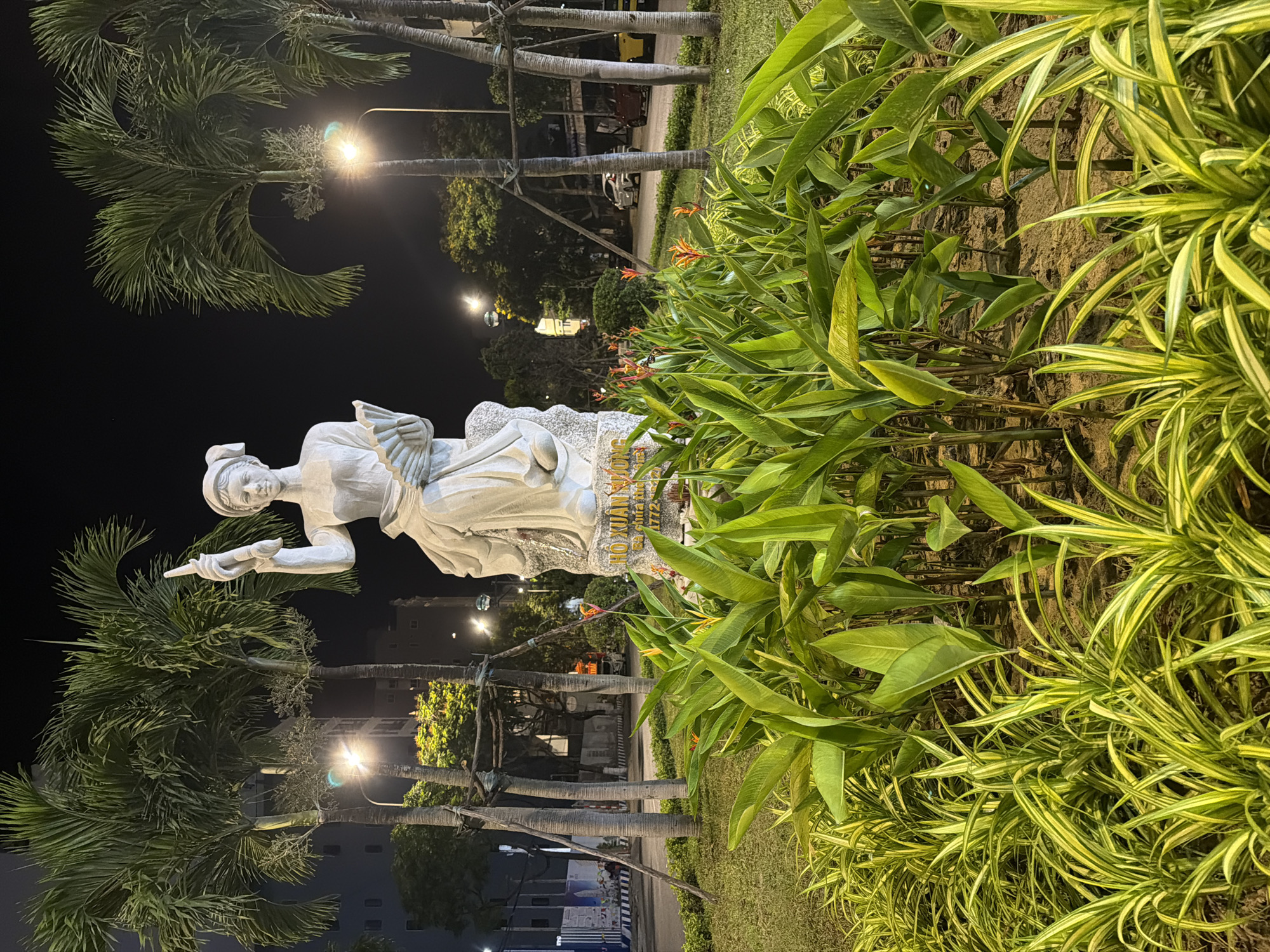 The image shows a statue of Ho Xuan Huong, a Vietnamese poetess, at night. The statue is located in Đà Nẵng, Vietnam. It is a white stone statue depicting a woman in traditional Vietnamese clothing, holding a writing instrument. The statue is surrounded by tropical plants, including palm trees and flowering bushes. Street lights illuminate the scene. In the background, buildings and vehicles are visible.