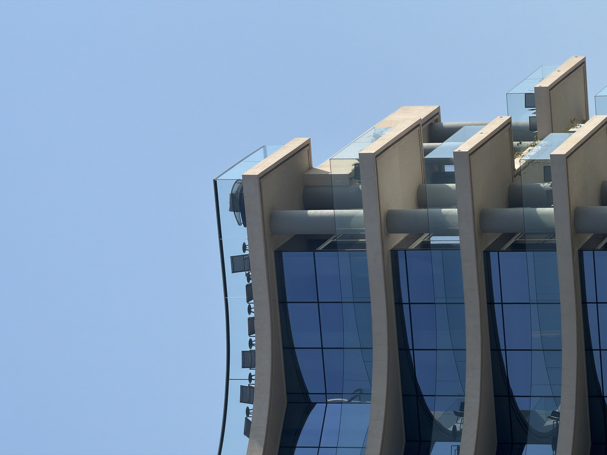 The image shows the exterior of a modern building with curved balconies against a clear blue sky. The building features multiple levels with large, dark blue reflective windows. Each balcony has a glass railing. On the uppermost balcony, several lounge chairs and small tables are visible. The architecture is characterized by smooth, light-colored concrete surfaces and cylindrical columns supporting the upper levels. The scene is brightly lit, suggesting daylight.