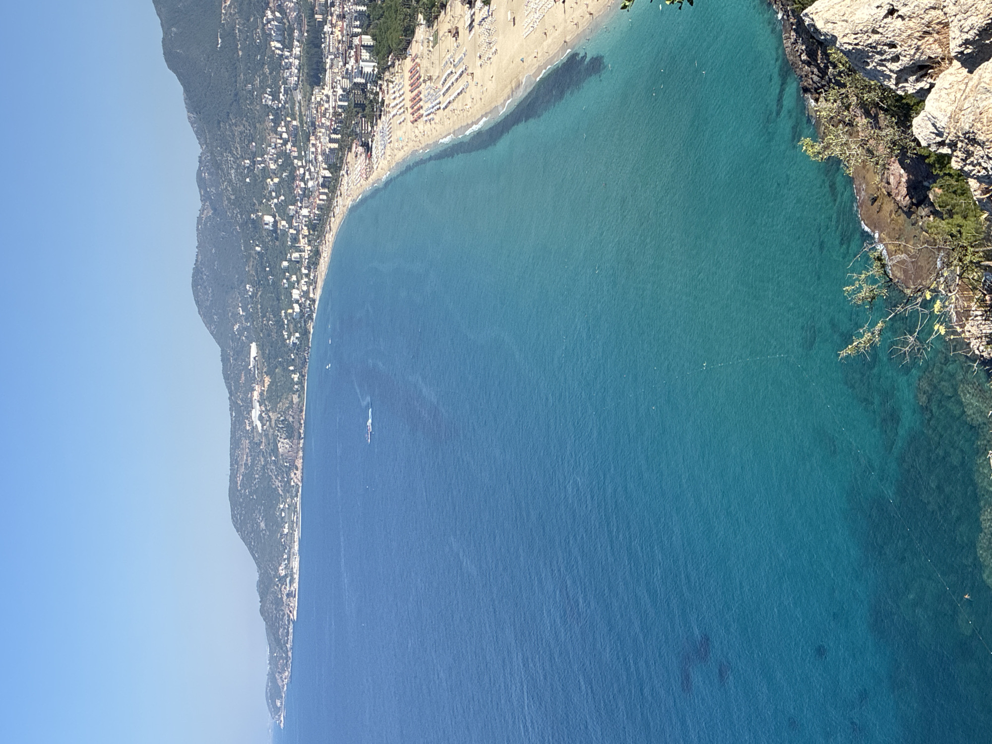 This is an aerial, high-angle, medium shot of the Cleopatra Beach in Alanya, Turkey, on a clear, sunny day. The turquoise water of the Mediterranean Sea dominates the left side of the frame, transitioning to a lighter blue closer to the shore. The beach, a crescent of light sand, is lined with numerous rows of white and orange beach chairs and umbrellas. A dense urban area, characterized by white and light-colored buildings, backs the beach, extending up the green, mountainous terrain. Trees, primarily green conifers, are interspersed within the city and along the slopes. Several small boats are visible on the water, and a few people can be seen on the beach and in the water. The foreground features a rocky outcrop with sparse vegetation.