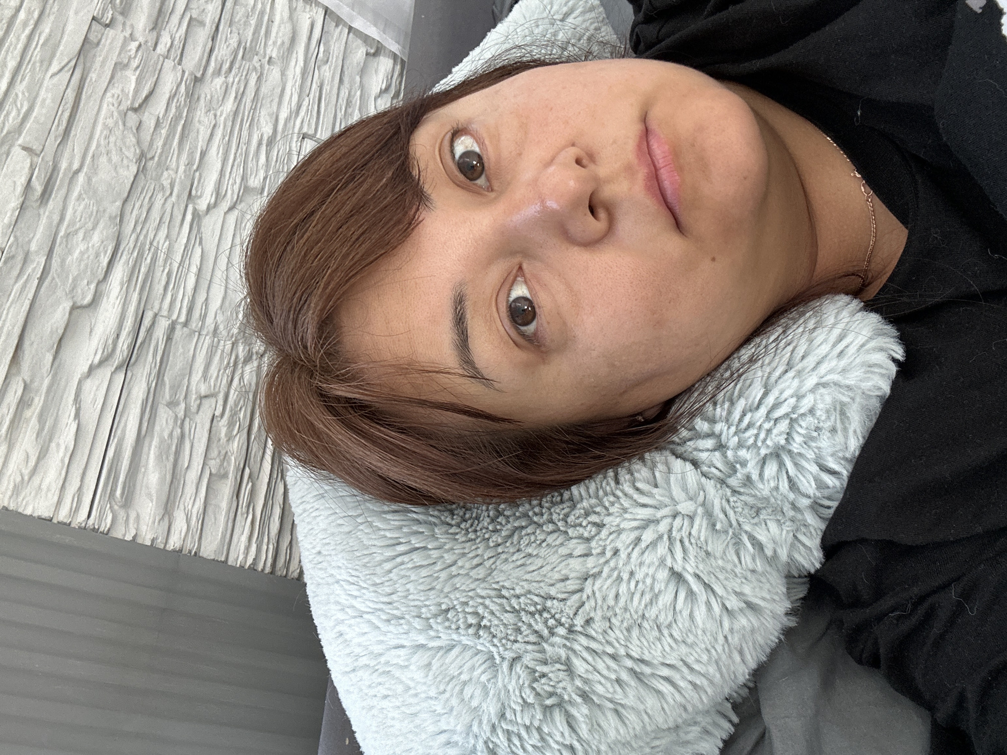 A person lies on a gray, furry pillow. They have brown hair and eyes. A black garment is visible, and a gold necklace is on the person's neck. A white, brick-like wall is in the background. The location is unknown.