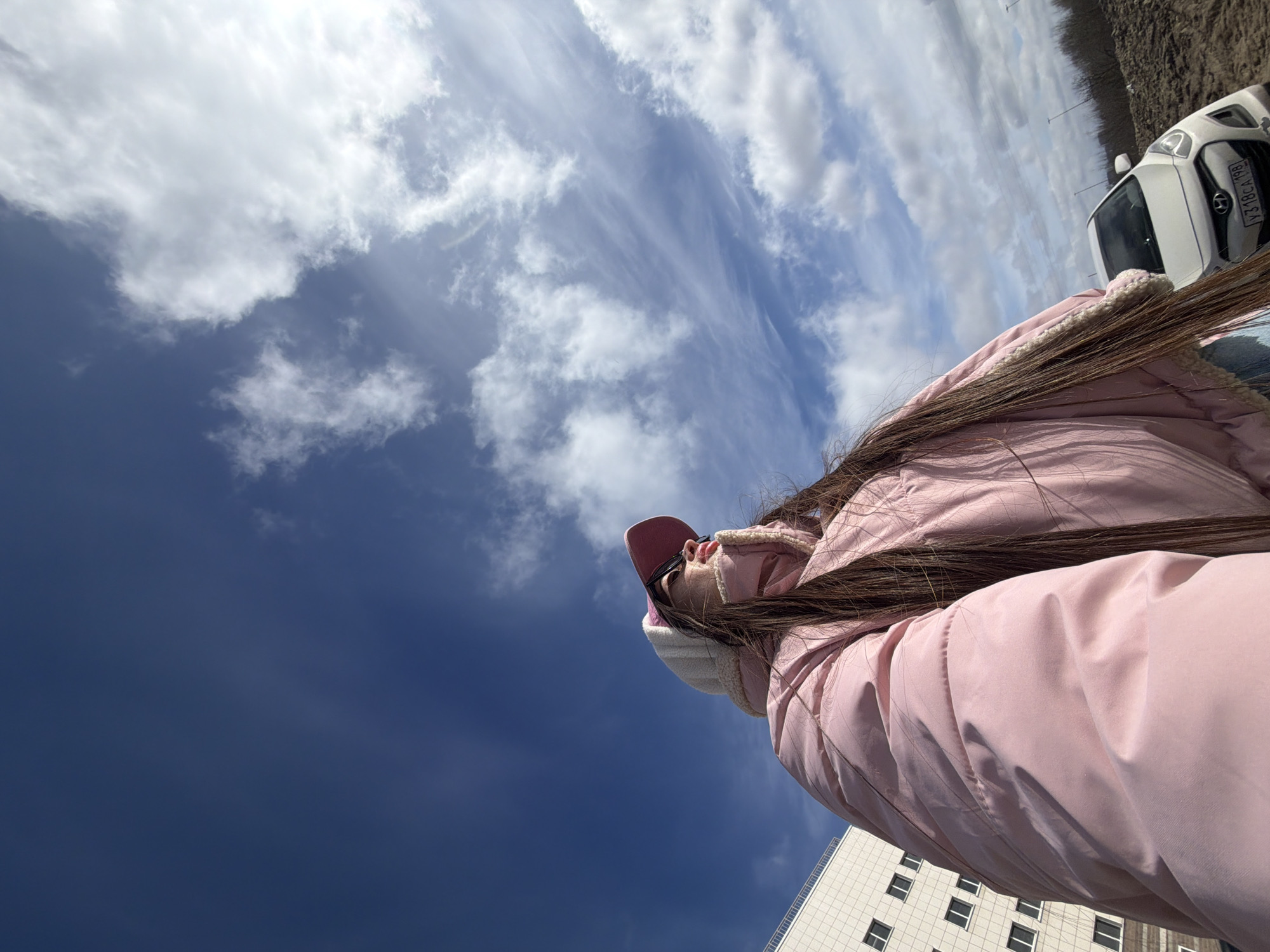 A low-angle, outdoor shot captures a person wearing a pink jacket and a pink cap. The person's long, dark hair partially obscures the view. Above, the sky transitions from a dark blue at the bottom to a brighter, cloud-filled sky at the top. A white car is visible in the upper right corner, along with what appears to be a grassy area and some trees. In the lower right corner, the corner of a light-colored building with multiple windows is visible. The location is possibly Saint Petersburg, Russia, considering the car plate.