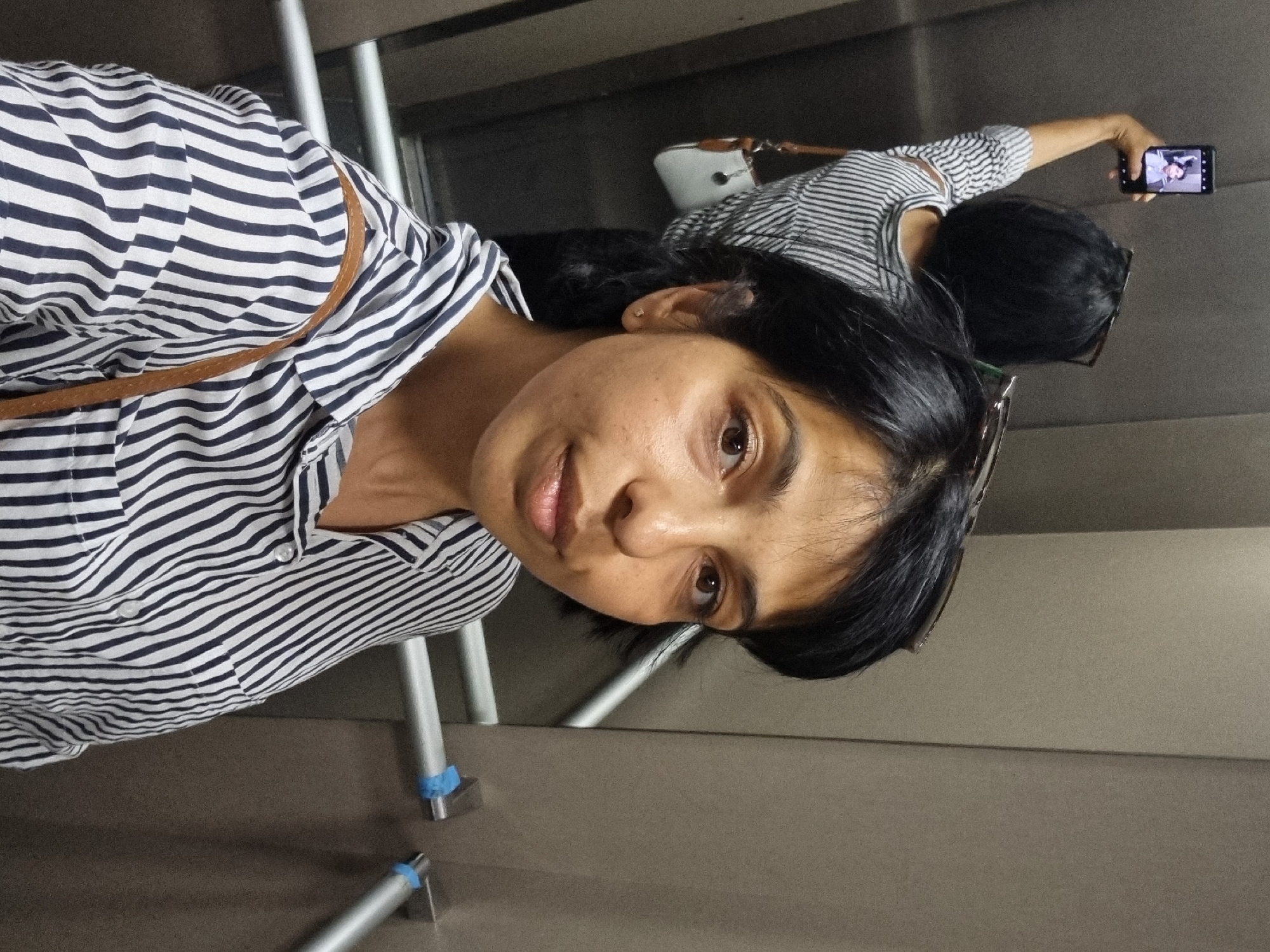 A woman is in an elevator, taking a selfie in the elevator mirror, while a second woman behind her is taking a photo of the first woman with her smartphone. Both women are wearing horizontally striped shirts. The elevator has a handrail made of metal tubing. The location of the photo is not possible to determine.