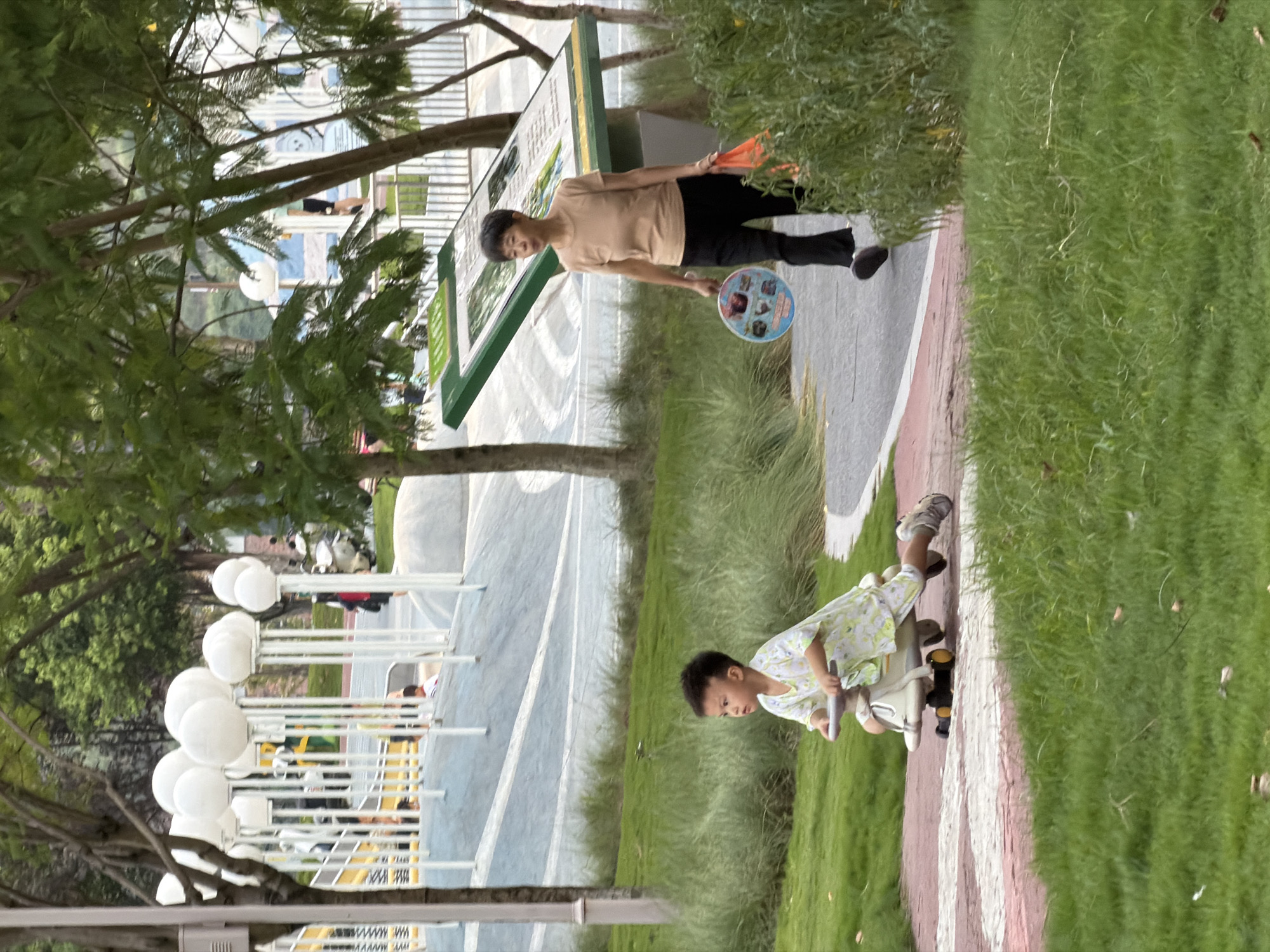 In Jiangjin, China, a young boy rides a toy vehicle down a park pathway. He is wearing a light-colored shirt with a leafy pattern and dark shorts. Ahead of him, a woman walks in the same direction, carrying a round fan in her left hand and an orange shopping bag in her right. She is wearing a tan short-sleeved shirt and black pants. The park features lush green grass, trees, and a playground with white spherical lights.