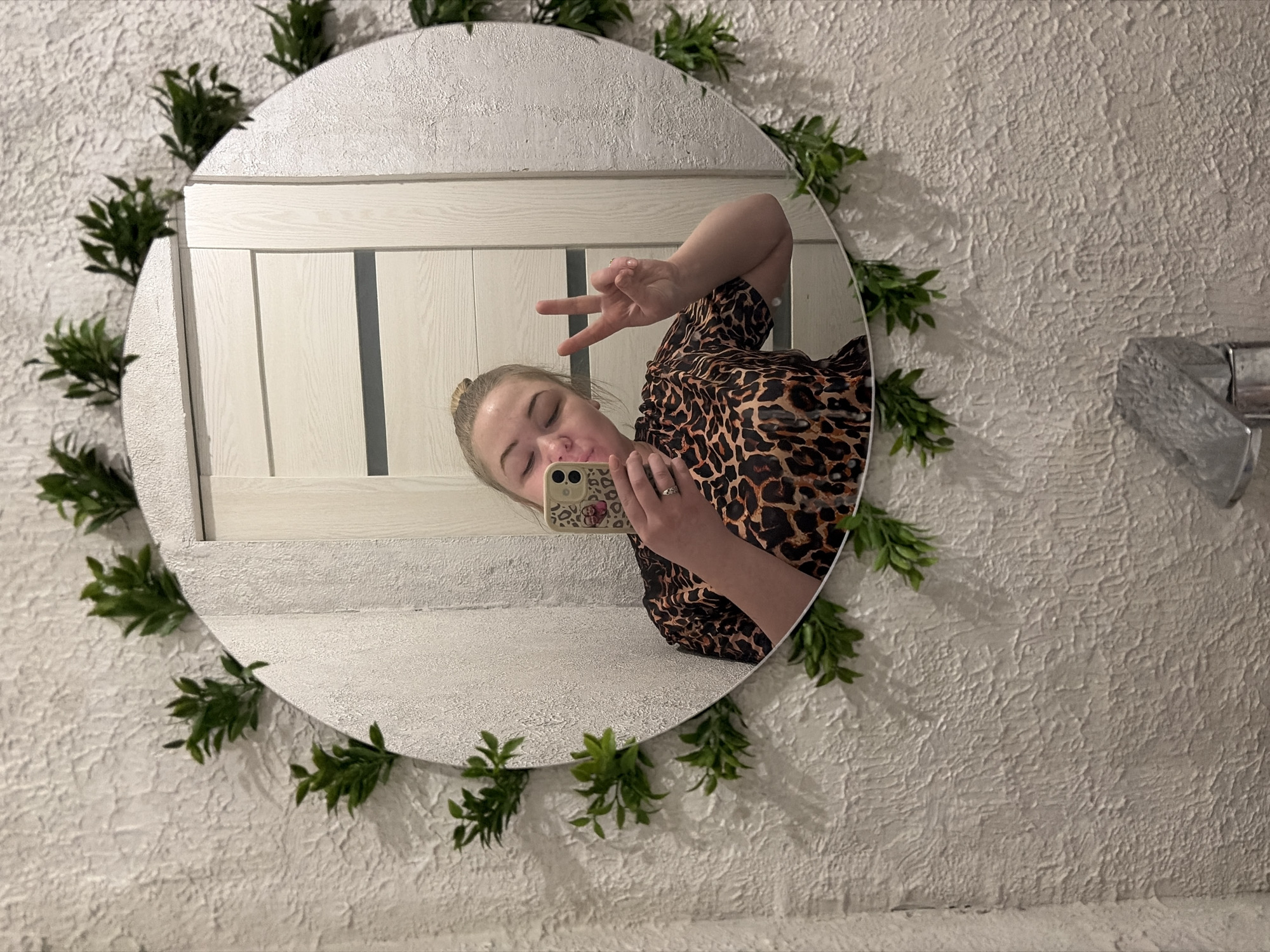 A young woman is depicted taking a mirror selfie in an indoor setting, likely in Noviy, Russia. She is reflected from the chest up in a round mirror, making a playful pose with her eyes closed or nearly closed, a puckered expression, and a "peace" sign gesture with her left hand. She wears a brown and black leopard print top or dress, and her light hair appears to be styled up. She holds a smartphone with a light-colored, patterned case featuring a small, unreadable graphic to capture the image.
The round mirror is decoratively framed by a garland of green faux foliage around its entire rim. The wall surrounding the mirror has a distinctive rough, textured finish and is off-white or light gray in color. Within the mirror's reflection, behind the woman, is a section of light-toned, possibly wooden, paneled furniture, suggestive of a headboard or cabinet. To the right of the mirror, a metallic, silver-colored faucet or fixture is partially visible on the textured wall, indicating a bathroom or similar washroom area. The lighting is artificial, and the overall mood conveyed by the woman's expression is one of lightheartedness and confidence. No readable text is present in the image.