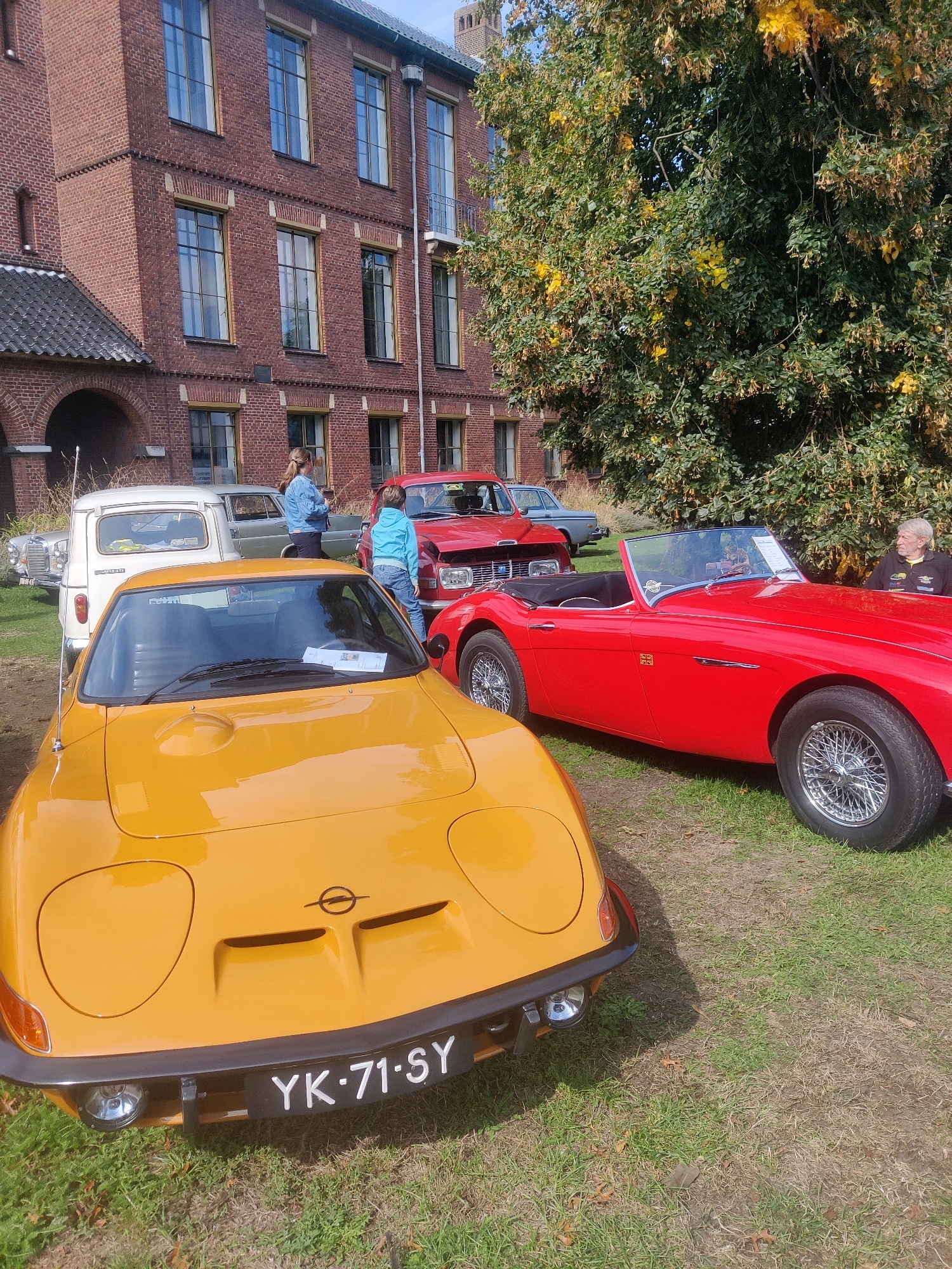 The image captures a sunny outdoor event, likely a car show, with several vintage vehicles parked on a grassy area in front of a red brick building. In the foreground, a bright yellow Opel GT sports car with its distinctive headlights and a license plate reading "YK-71-SY" is prominently displayed. To its right, a vibrant red convertible, possibly a classic British roadster, features chrome wire wheels and a Union Jack emblem on its fender. Further back, a white Renault 4 van and other cars are visible. Two women, one in a blue jacket and the other in a light blue hooded sweatshirt, are standing near the vehicles. An older man with gray hair is seated to the right of the red convertible, wearing a dark jacket with yellow accents. The building in the background appears to be an institutional or historic structure with large, multi-paned windows. Lush green trees with some yellowing autumn foliage provide a natural backdrop. The overall scene suggests a casual gathering focused on classic automobiles, set in a location that is identified as the city of Winterswijk, Netherlands.