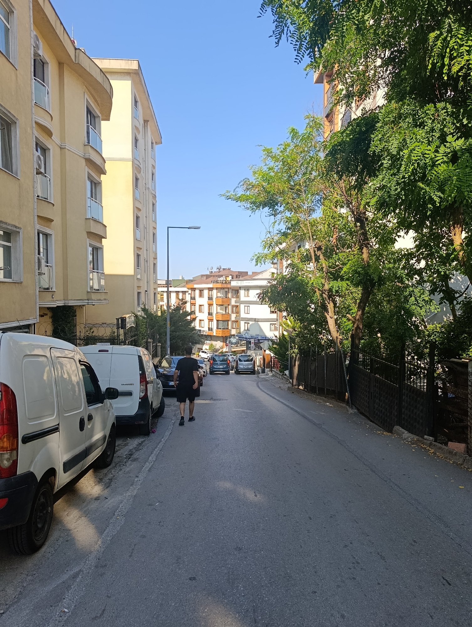 The image shows a street scene in Ümraniye, Turkey. On the left side of the street, there are two yellow apartment buildings with multiple balconies and windows. Parked along the curb are two white vans and other vehicles. A person wearing a black t-shirt and shorts is walking away from the camera down the street. The street itself is paved and slopes slightly upwards. On the right side of the street, there are trees with lush green foliage, a black metal fence, and apartment buildings in the distance. The sky is clear and blue.