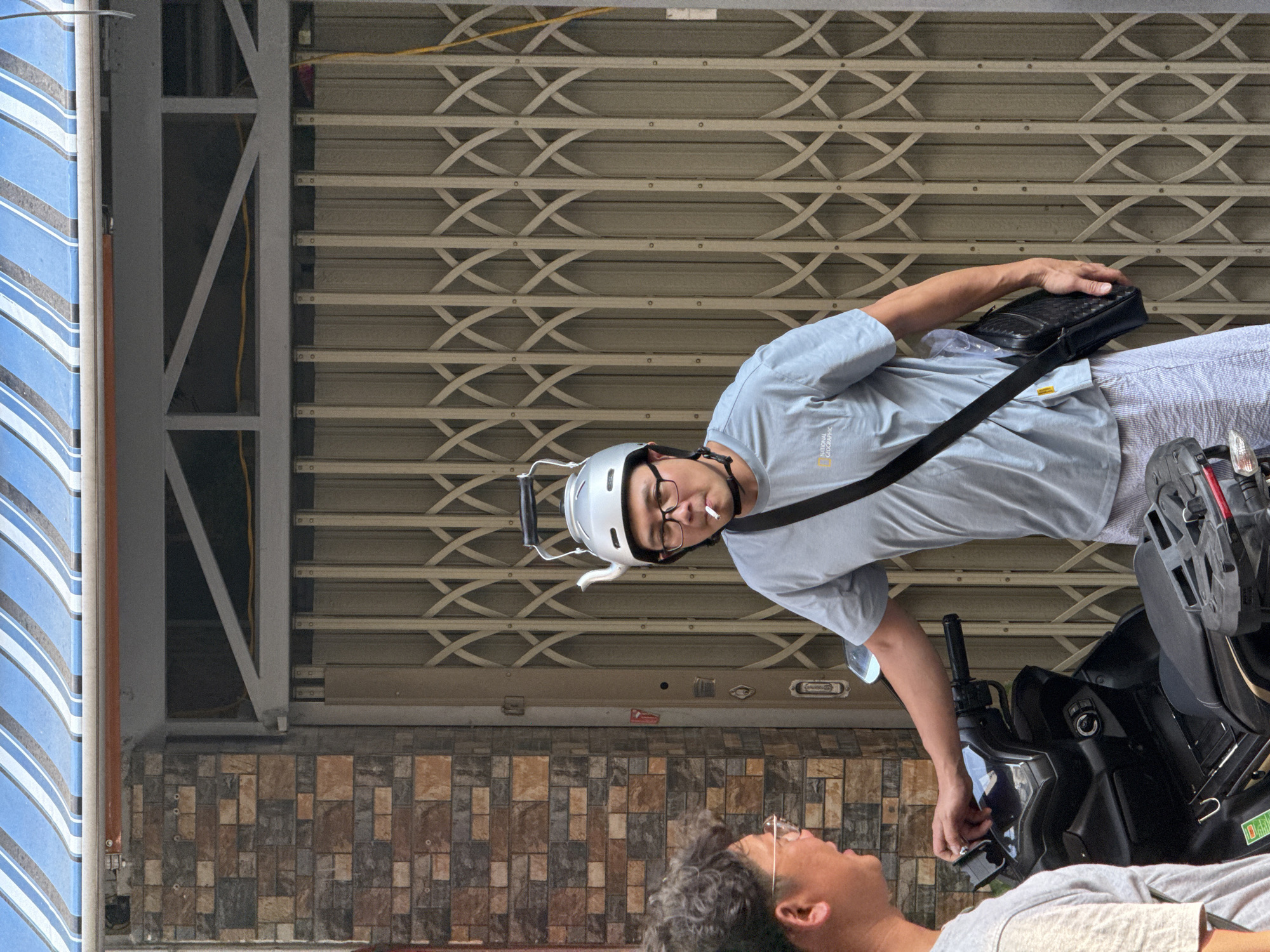 A medium, slightly rotated shot captures two Asian men interacting in front of a closed storefront with a patterned metal rolling gate. The ground is covered with multi-colored brick patterns. The storefront is located in Nha Trang Ward, Vietnam.
One man, wearing a light blue shirt, light-colored striped pants, and a silver helmet with a smoking cigarette in his mouth, looks directly at the camera. He is holding a black bag with a shoulder strap. The second man, wearing a light gray shirt, looks up at the first. A black moped is between the two men.
In the background, a blue and white striped awning is partially visible, along with a metal support structure.