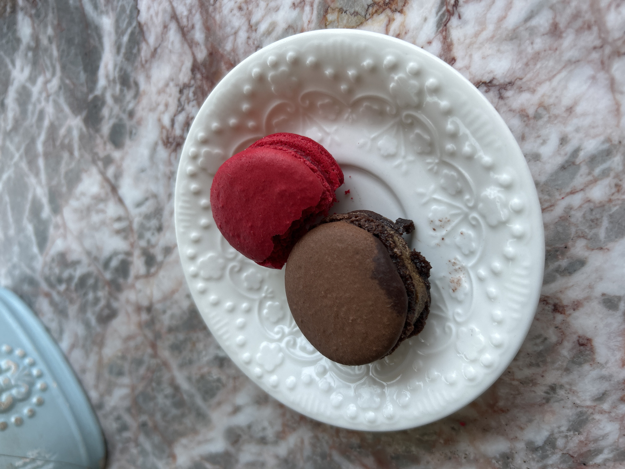 An overhead, close-up shot captures two macarons on a white, ornate ceramic plate, set against a marbled background. The plate features a scalloped edge adorned with raised dots and intricate, swirling floral-like patterns. One macaron is a vibrant red, with a noticeable bite taken out, revealing its soft, textured filling. Beside it rests a dark brown macaron, possibly chocolate, which also appears partially eaten or has a crumbly edge.
The background is a polished marble surface, displaying a natural pattern of white, grey, and reddish-brown veins, suggesting a countertop or table. In the bottom-left corner of the frame, a segment of a light blue object with similar embossed decorative elements as the white plate is visible. The lighting is bright and even, indicating an indoor setting during the daytime. The image evokes a sense of sweet indulgence and a moment of quiet enjoyment. There is no visible text in the image.