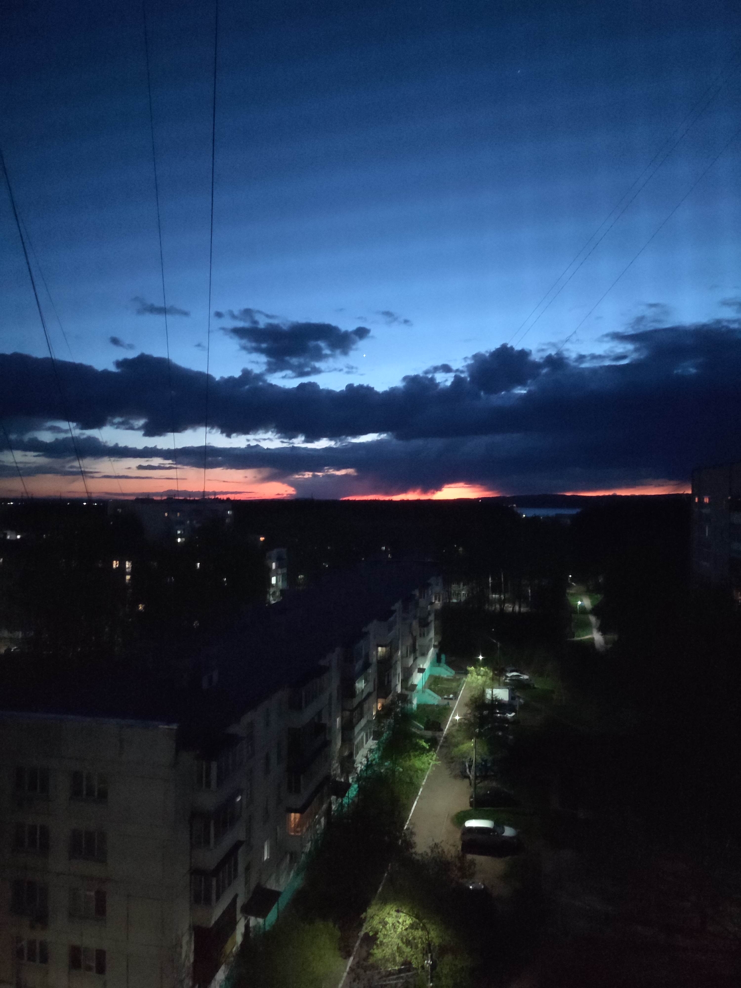 A nighttime cityscape under a twilight sky. The sky transitions from a deep blue to a reddish-orange on the horizon, with dark clouds scattered across. Silhouetted buildings are visible in the middle ground, with sparse light emanating from some windows. A residential street with trees, parked cars, and lit street lamps runs along the base of the buildings. Power lines are visible in the upper left corner of the frame. The location is the city of Unknown, Unknown.