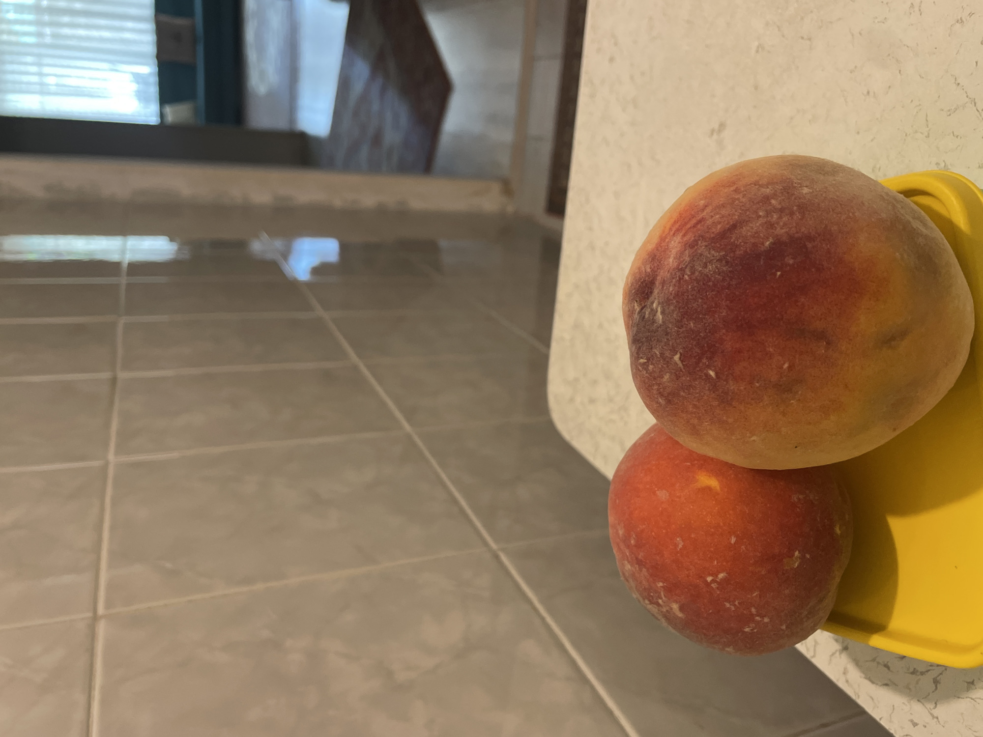 Two ripe peaches are shown on a yellow dish in the foreground, positioned on a speckled countertop. Behind the peaches, a tiled wall occupies the left side of the image, with the tiled pattern angled upwards. To the right, an open doorway reveals a glimpse of a room with dark flooring, a rug, and a sofa with cushions under a window with sheer curtains. The scene appears to be in a kitchen or dining area. There are no people present. The lighting is natural and coming from the right side of the frame. The overall impression is domestic and casual. There is no information in the image to suggest the location is Kadıköy, Turkey.