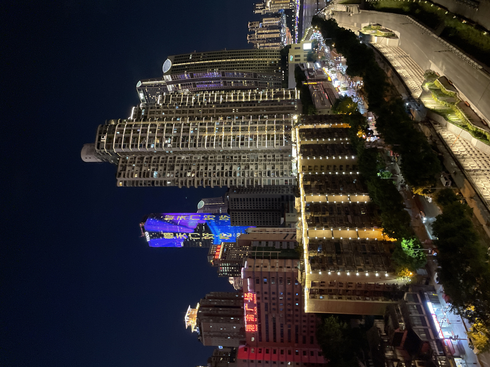 The image depicts a nighttime view of a dense urban landscape in Yuzhong District, China. The foreground features a multi-story building with illuminated windows and vertical light strips. To the right, a sloping road with white lines is visible, flanked by a terraced wall covered in greenery and illuminated by artificial lights. Further into the background, a cluster of modern high-rise buildings dominate the skyline, many of them illuminated with colorful lights and signage. One prominent skyscraper features a bright blue facade with Chinese characters displayed in yellow light, and the word "VOCO" in white. Another tall building showcases a grid-like pattern of lights, highlighting its structure. The overall scene suggests a vibrant and active city at night, with a mix of residential and commercial architecture.