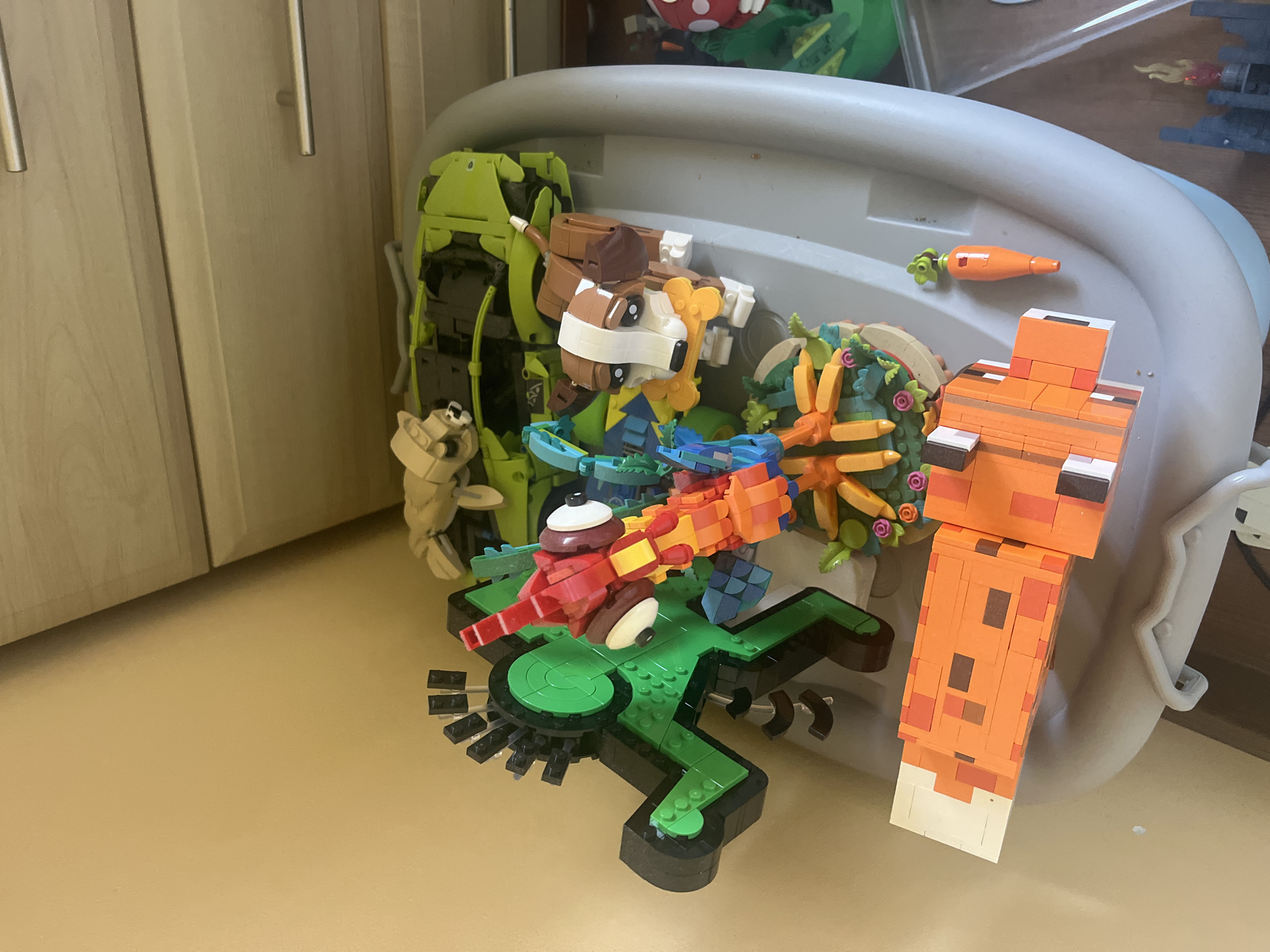 The image displays an indoor scene featuring a collection of various Lego models arranged on a light grey, contoured plastic surface, possibly a high chair tray or a large storage container lid. The background consists of a plain beige-colored wall on the left and a light wooden dresser with two silver horizontal pull handles on the right. The location of the scene, including any specific city such as Onokivtsi, Ukraine, is not discernible from the provided image.
Prominently featured on the left is a large, stylized Lego figure, primarily green and black, with a spiky black headpiece and large circular green elements on its body. To its right, another colorful Lego creature, predominantly red, orange, and yellow, with large white eyes and a beak-like mouth, is displayed. Below this creature, a small, circular Lego base holds various orange, green, and pink elements arranged to resemble plants or flora.
Towards the center-right, a brown and white Lego dog figure, resembling a beagle, is depicted holding a yellow bone in its mouth. Behind the dog, a bright lime-green Lego sports car is partially visible, appearing to be on its side or upside down. A smaller, tan-colored Lego creature is also visible resting near the top right of the green car.
In the foreground, a large, blocky orange Lego fox figure, designed in a pixelated style, lies flat on the grey surface. A small, orange Lego carrot with a green top is positioned next to the fox. A partial view of additional Lego constructions, including a dark grey structure with a red flame element and a red and white spotted mushroom, is visible in the bottom right corner. No people are present in the image, and the objects are static, arranged for display.