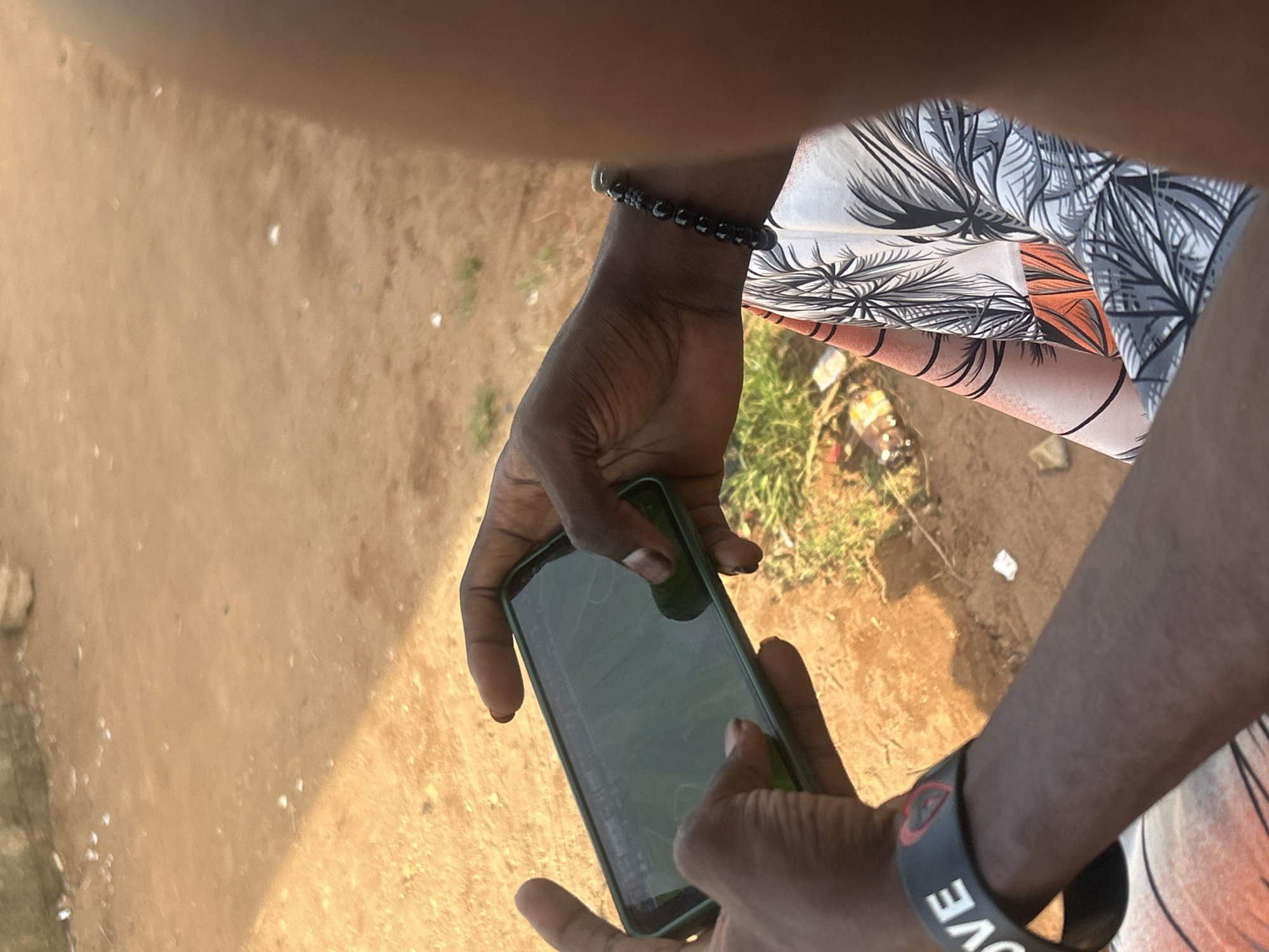 A person holds a cell phone with a green case. The person is wearing a bracelet with the word "LOVE" on the wrist, and another bracelet of beads on the opposite wrist. The person is wearing a shirt with a palm frond pattern in black and white with orange. The ground is dirt and grass. The location is Unknown, Unknown.