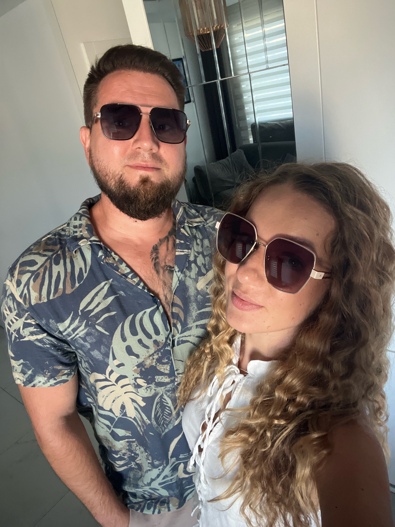 The image depicts a couple taking a selfie, standing indoors, likely in a residential setting. The man on the left is wearing a short-sleeved, blue, tropical-print shirt with large leaves and a beard, and sunglasses. He has tattoos visible on his chest. The woman on the right is wearing a white, ruffled top and also has sunglasses on. Her long, curly, blonde hair frames her face. The background shows a mirrored wardrobe with blinds behind it, and a glimpse of a couch. The lighting suggests it is daytime. There is no specific indication in the image that places them in Alanya, Turkey.
