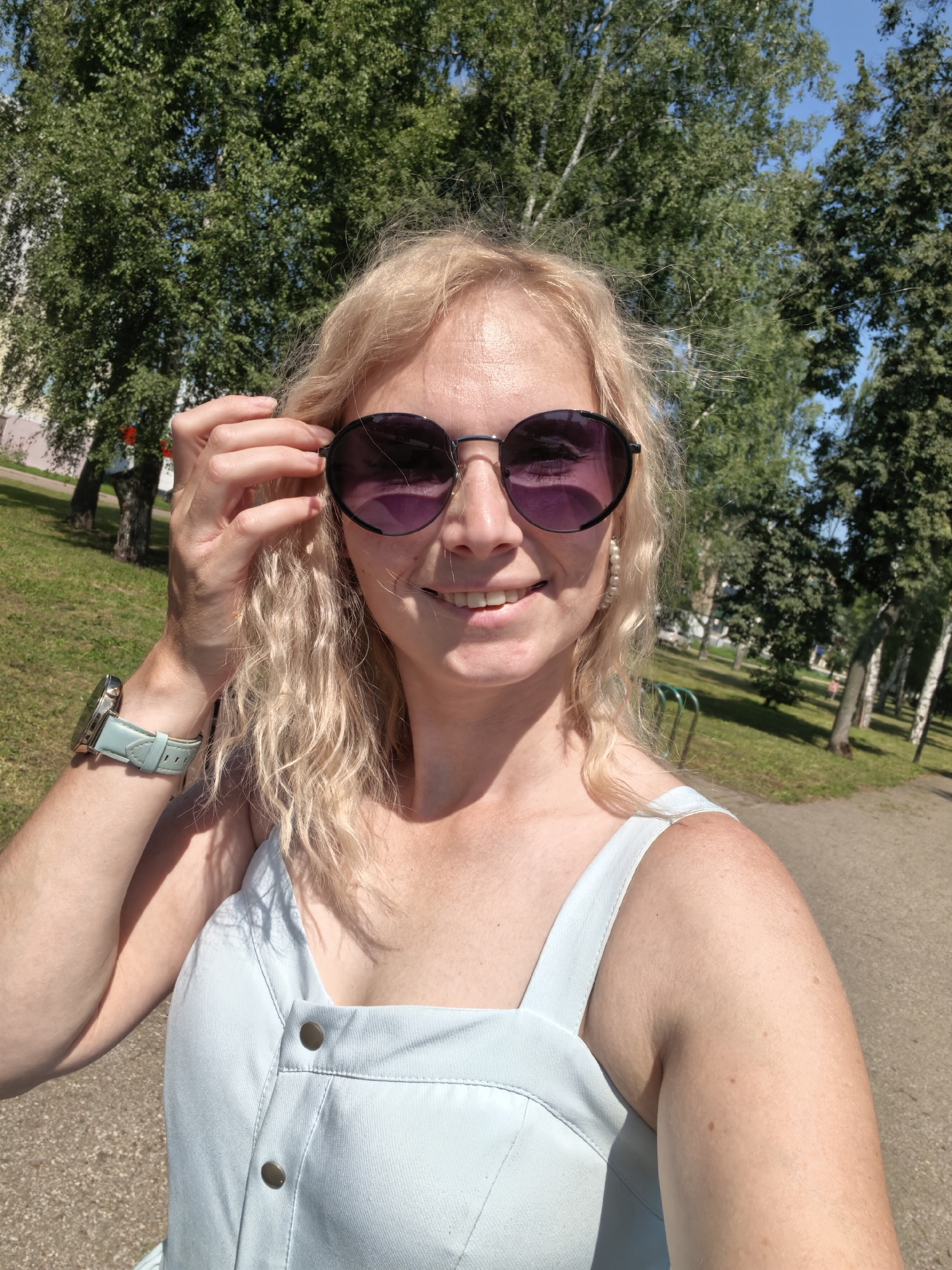 A medium-shot selfie shows a woman with blonde, wavy hair and sunglasses in an outdoor park setting. She is wearing a light blue dress and a light blue watch on her left wrist. The background consists of green trees and a paved walkway, suggesting a sunny day. The location is identified as the city of Sterlitamak, Russia.