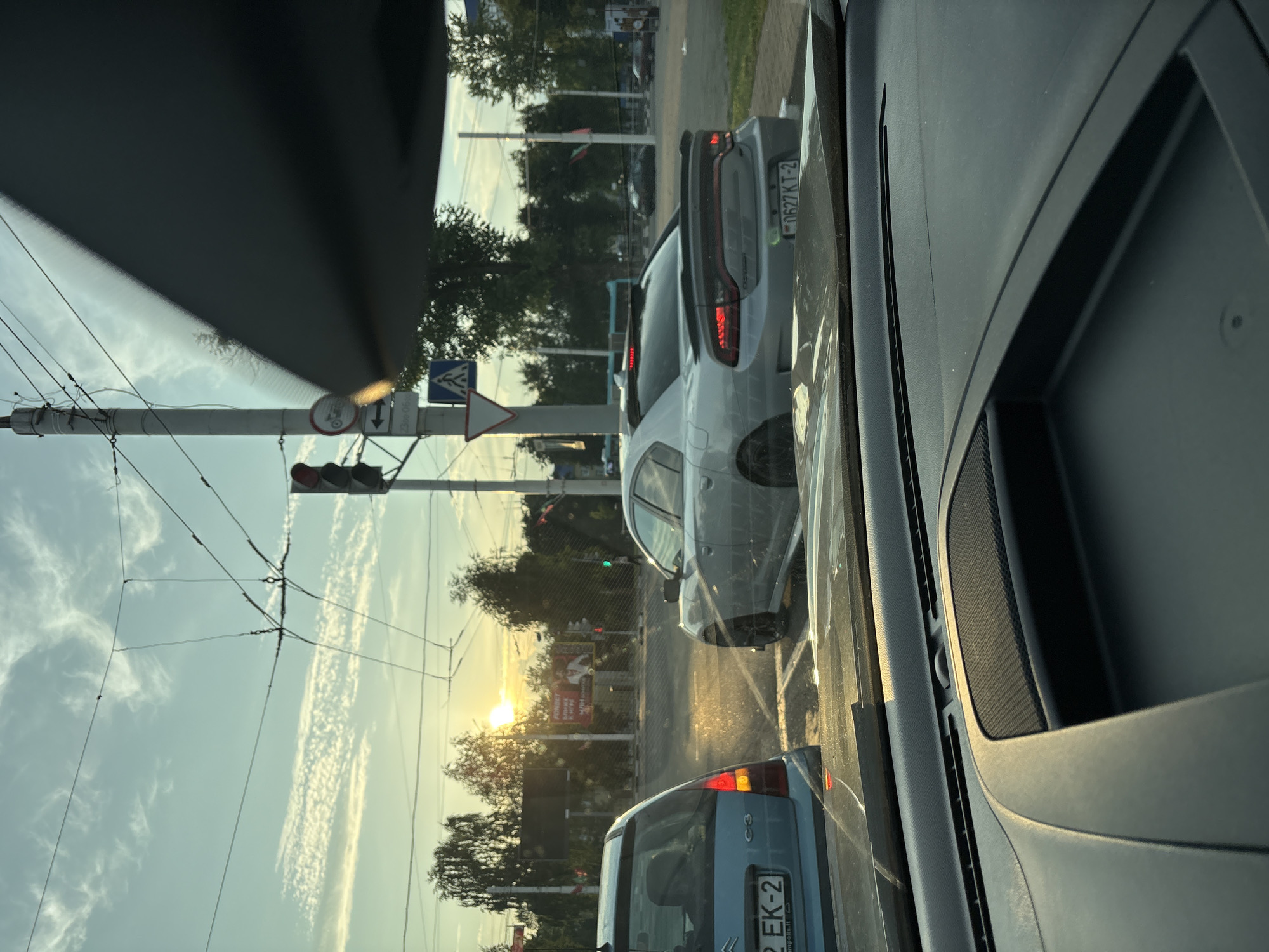 This is a photograph taken from inside a vehicle, looking out at an intersection in Vitebsk, Belarus. The sun is low in the sky, casting a warm glow and creating lens flare on the windshield. In the foreground, the interior of the car is visible, including the dashboard and a portion of the passenger seat.
The intersection features traffic lights, overhead wires for what appears to be a tram or trolleybus system, and various road signs. Two cars are stopped at the intersection: a light blue Citroën C3 on the left with its license plate partially visible, and a silver Dodge Charger on the right with its rear brake lights illuminated. The Charger's license plate is also partially visible. Trees line the sides of the road, and a billboard advertising "FONBET" is visible in the background. The sky is partially cloudy with wispy cirrus clouds. The overall impression is a typical urban street scene during sunset.
