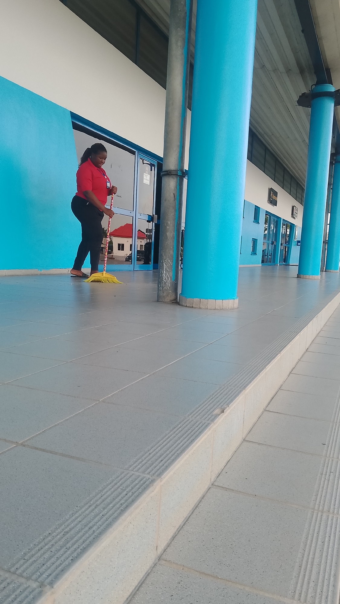 A woman in a red shirt and black pants is sweeping a tiled floor with a yellow broom. She is standing in front of a building with blue walls and columns. The building has glass doors and windows, and a red-roofed building is reflected in one of the glass doors. The scene is on a walkway or patio in the city of Orerokpe, Nigeria.