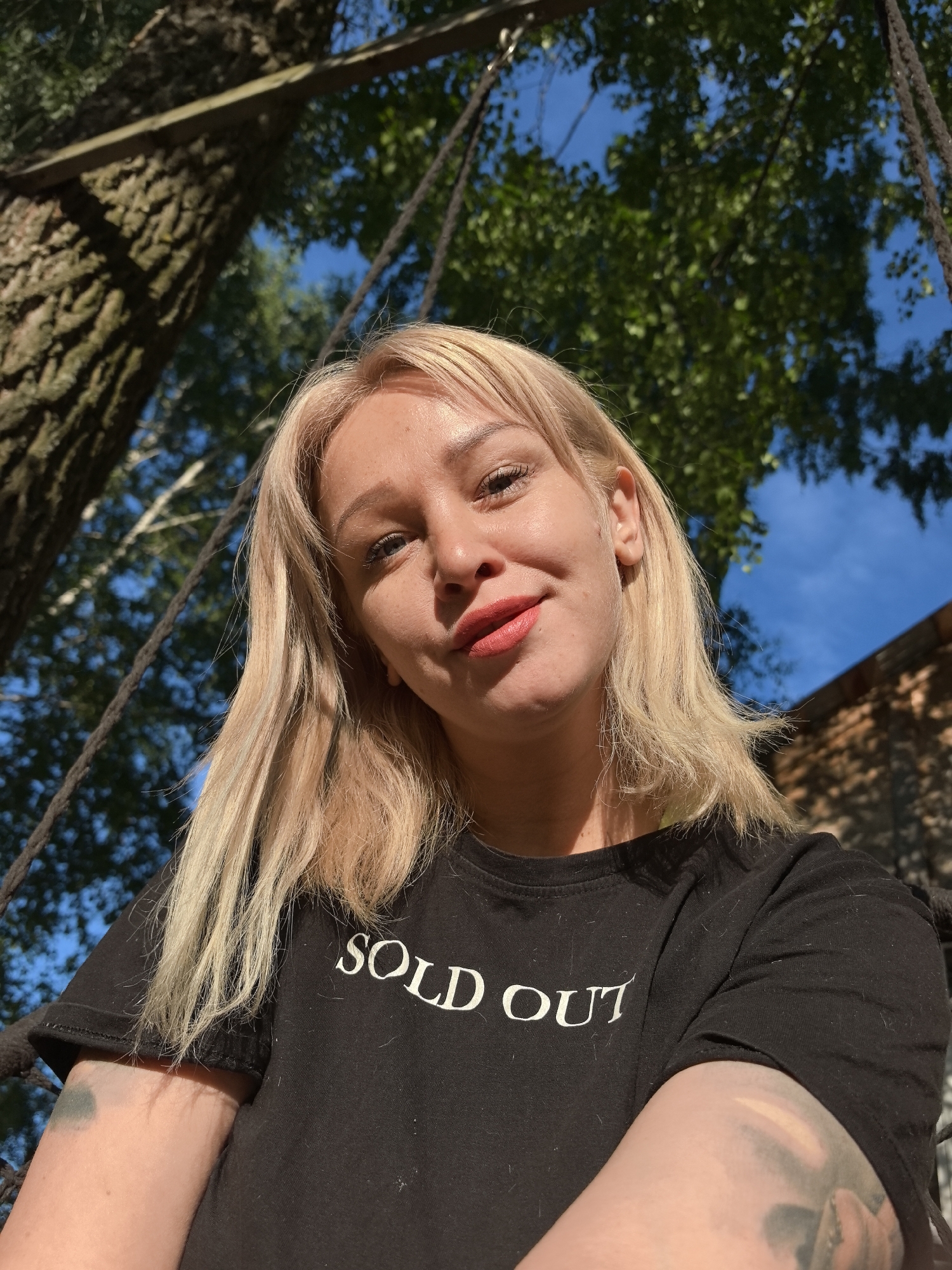 A young adult female, estimated between 25-35 years of age, is depicted in a close-up, upward-angled outdoor selfie photograph taken in Noviy, Russia. She has blonde, shoulder-length hair with a fringe, and light-colored eyes. Her lips are adorned with red/coral lipstick, and she exhibits a slight smirk. She wears a black short-sleeved t-shirt with the white text "SOLD OUT" centrally printed on the chest. Her right forearm features a distinct dark green/blue circular tattoo, and her left forearm shows part of a larger, indistinct dark tattoo. Dark, braided ropes are visible on either side of the subject, indicating she is likely seated on a swing or hammock. The background features a clear blue sky and abundant green foliage from deciduous trees, suggesting a sunny outdoor environment. A large tree trunk with rough bark is prominent on the left, with a horizontal wooden beam or plank traversing near the top left. A sunlit stone or brick wall and a roofline of a building are partially visible on the far right. Strong natural sunlight illuminates the scene, casting shadows on the subject's face and highlighting the background.