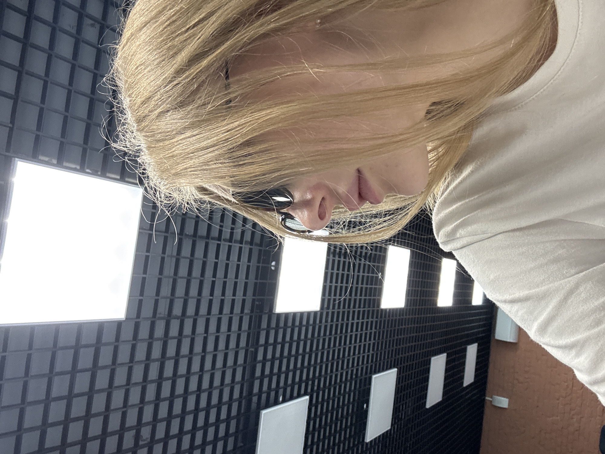 The image features a woman with blonde hair wearing glasses and a white shirt. The background consists of a dark grey tiled wall with square cutouts, some of which are lit, possibly from behind. The scene appears to be indoors.
