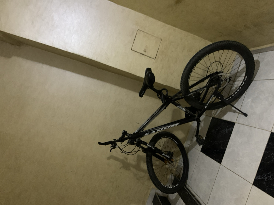 A black mountain bike leans against a beige wall and checkered floor. The bike's rear wheel is slightly elevated, with a kickstand visible. The front wheel faces the wall. The bicycle's frame has visible lettering. A small, rectangular access panel is on the upper wall near the bike. The lighting is somewhat dim, creating soft shadows around the bike and on the textured wall.
