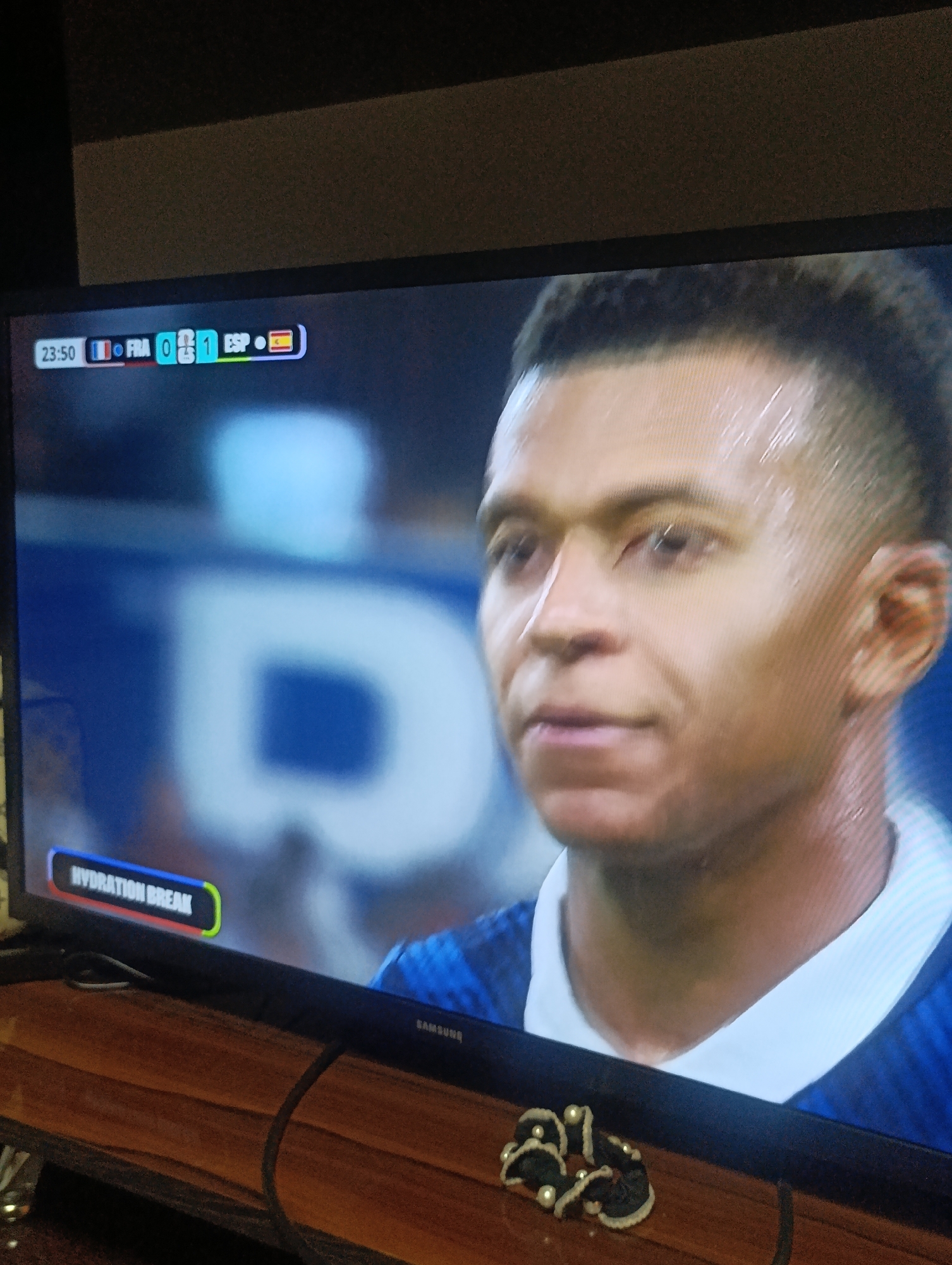A Samsung television screen displays a football match with the score "FRA 0-1 ESP" at 23:50. The screen features a close-up of a player, likely from the French team given the score, with sweat visible on his forehead and neck. The background shows large, blurred white lettering. In the bottom left corner, a small graphic reads "HYDRATION BREAK." The television is placed on a wooden surface, and a dark cable snakes across the screen and down the front. The overall setting appears to be indoors. There is no indication that this scene is located in Osh City, Kyrgyzstan.