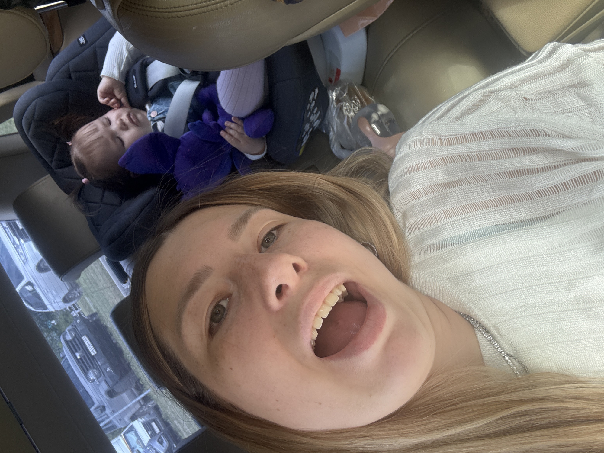 A selfie taken inside a vehicle features a woman with light brown hair, light skin, and freckles, wearing a white, textured shirt and a silver necklace, with her mouth open wide. Behind her, a child sits securely in a car seat, clutching a purple stuffed toy. The child is also wearing a white shirt and appears to be asleep or resting. Part of the vehicle's interior, including the leather seats and roof, is visible. Through the vehicle's windows, various parked vehicles and greenery suggest an outdoor environment. The registration plates on the vehicles indicate the location might be Moscow, Russia.