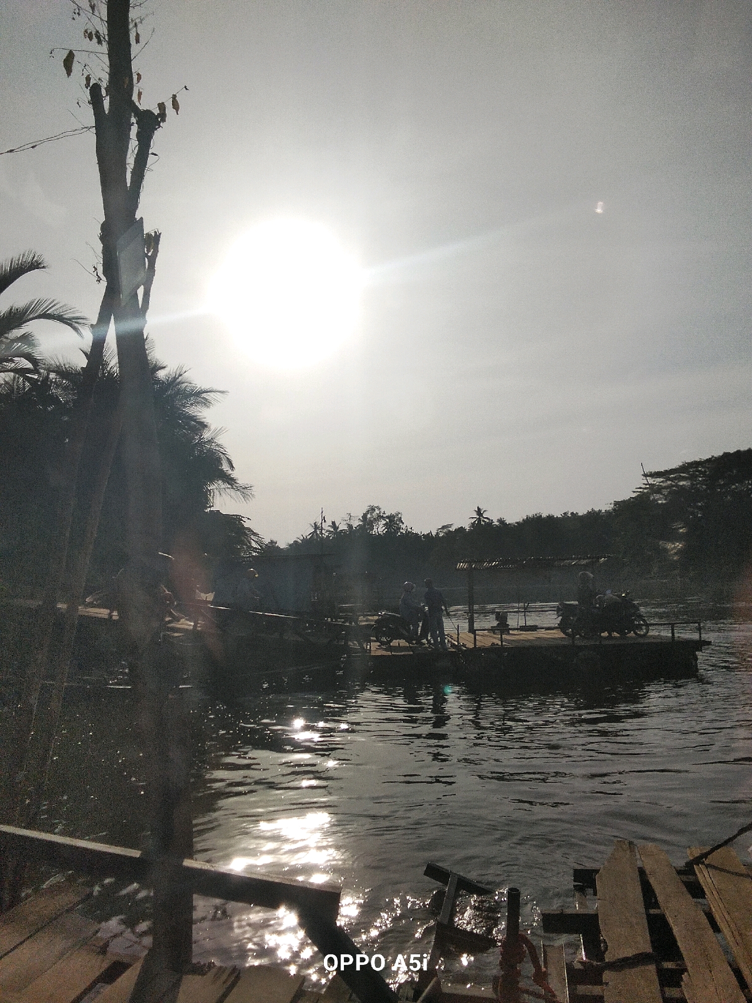 A wide-angle, low-light, outdoor scene depicts a tranquil river scene in Tabanggele, Indonesia. The sun, partially obscured by haze, shines brightly in the upper left quadrant, casting a strong glare. In the foreground, a wooden dock is visible, with planks of wood laid out. The river's surface reflects the sunlight, creating shimmering patterns. A wooden structure, possibly a raft or a small boat, floats in the middle ground, carrying several people and two motorcycles. The people appear to be engaged in conversation or preparing to depart. The background is a dense line of trees and vegetation, with a few distant structures and utility poles. The overall atmosphere is one of a peaceful, rural setting at sunset.