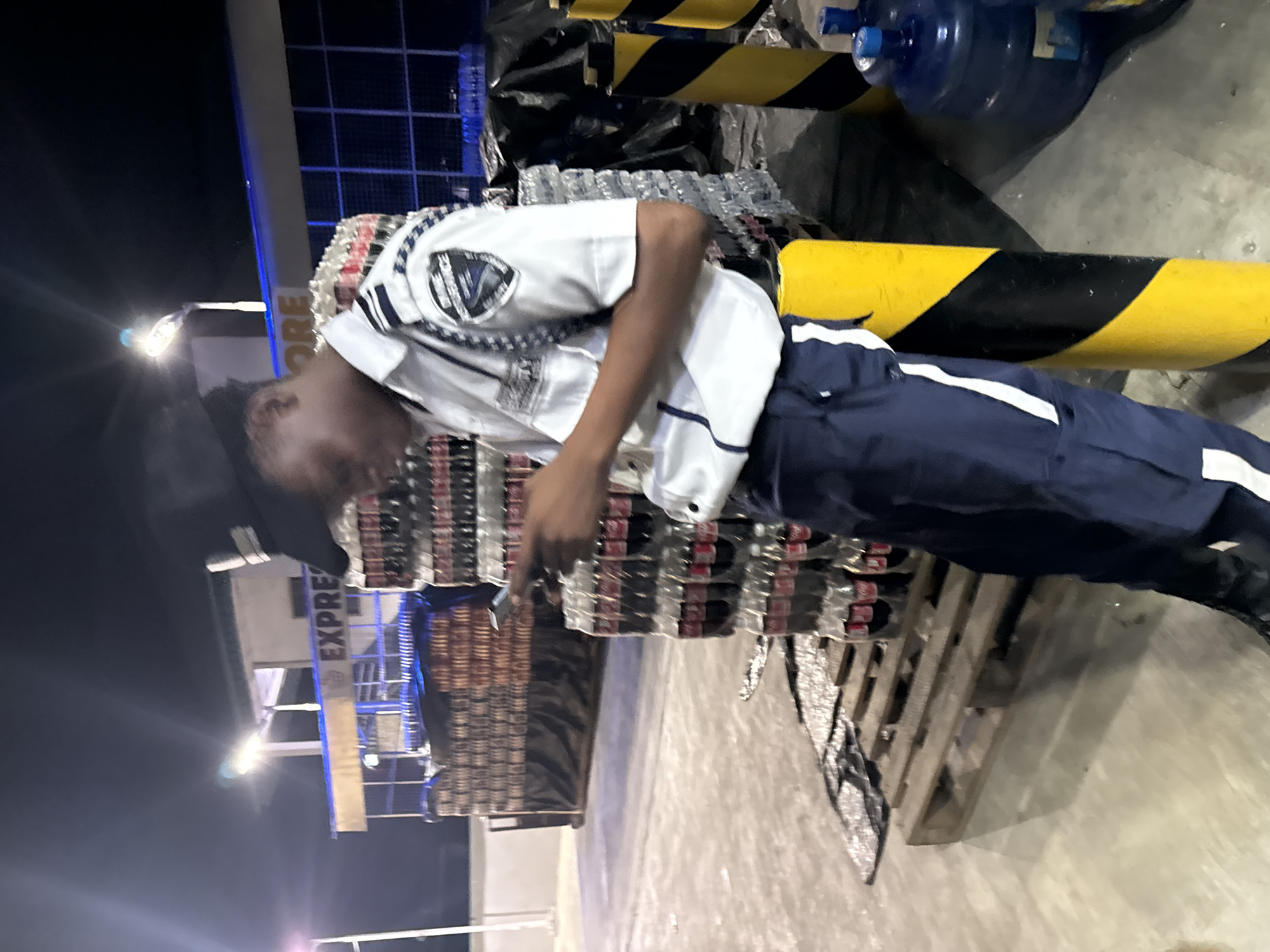 The image displays a nighttime outdoor scene, likely a commercial or storage area. A male individual, appearing to be a security guard, is positioned slightly right of the frame's center. He wears a white short-sleeved uniform shirt with a dark emblem on the left shoulder, which partially reveals text including "DIVINE" and "GOVERNANCE." He also wears dark blue trousers with a vertical white stripe on the outer seam and a dark cap. The individual is looking down at a smartphone, which he holds with both hands. His body leans against a yellow and black striped cylindrical bollard to his right.
To the left and extending into the background, numerous large stacks of bottled beverages, identifiable as Coca-Cola products, are arranged on wooden pallets. Further into the background, additional stacks of bottled water, some partially covered with black tarps, are visible. The ground is a light-colored paved surface, exhibiting some reflective properties. In the mid-ground to the right, a large blue water dispenser bottle rests on the ground near another yellow and black striped bollard.
In the far background, a building structure is illuminated by artificial lights, featuring a sign with the partially visible text "EXPRESS." Multiple bright artificial light sources are present, creating noticeable lens flares against the dark night sky. No explicit visual information within the image confirms the location as the city of Ado, Nigeria.