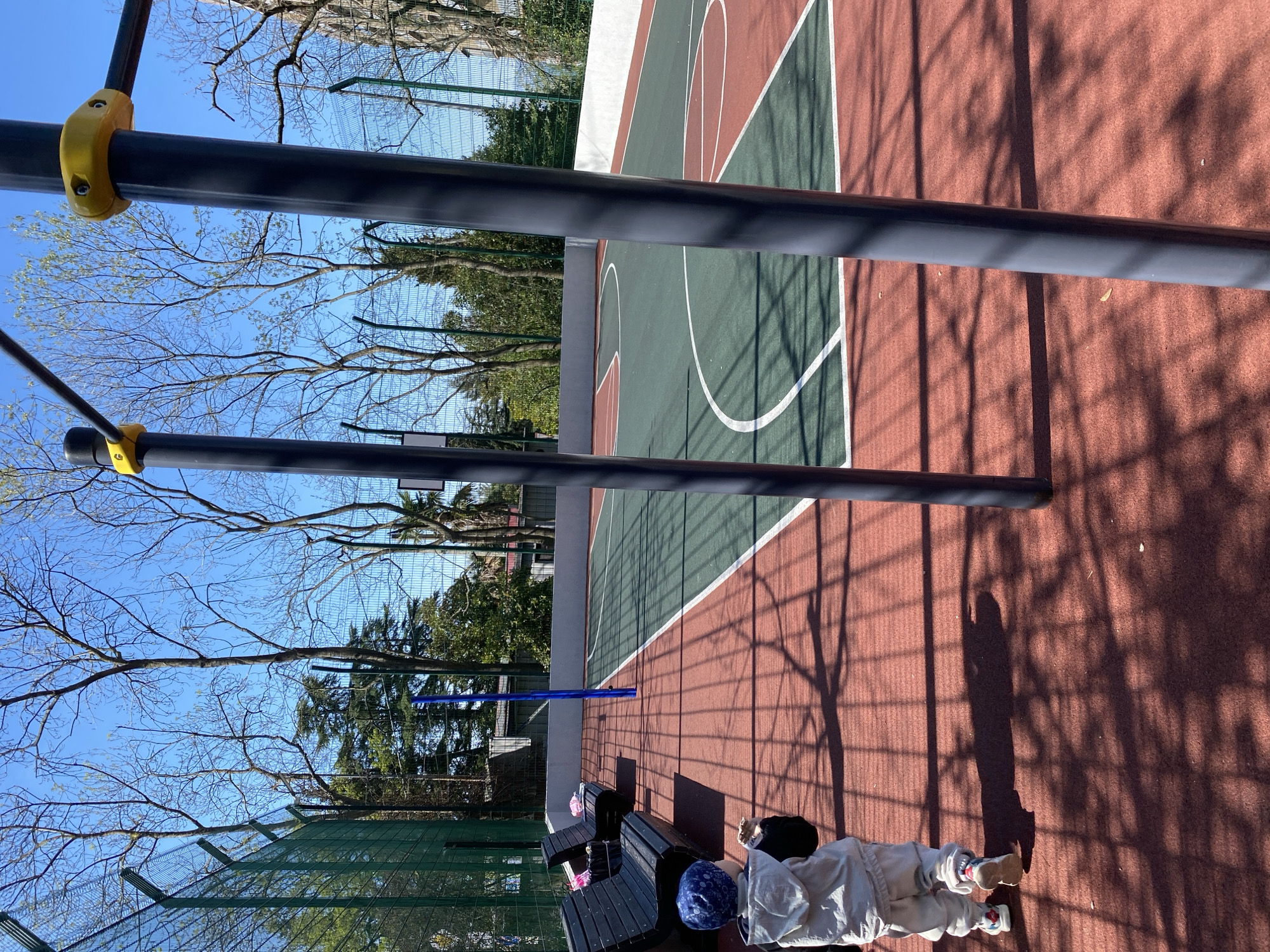 The image depicts an outdoor scene at a sports facility in Sochi, Russia. A green and brown basketball court is visible, with standard court markings. Two horizontal black bars, part of exercise equipment, are in the foreground. The shadow of a fence is cast on the court. To the side of the court, there are wooden benches and an individual lying on the ground near them. The person is wearing light-colored clothing, a blue patterned hat, and shoes. Trees and a chain-link fence surround the court. The sky is blue with bare tree branches.