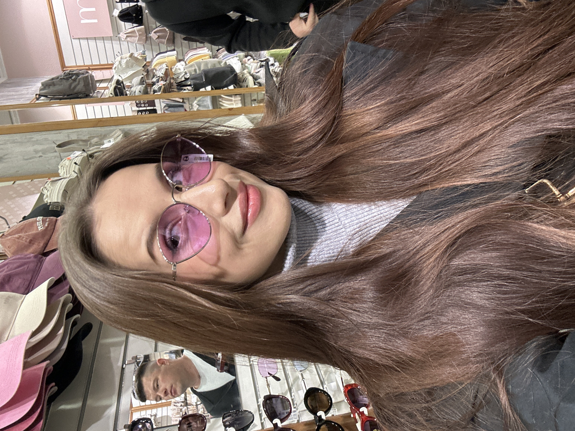 A woman with long brown hair is wearing pink sunglasses and a gray turtleneck shirt. She is indoors, possibly in a clothing store or boutique. Various items such as hats, bags, and clothing are displayed on shelves and racks in the background. The reflection of a man can be seen in a mirror. There is no information in the image to support the assumption of the location in the city of Yekaterinburg, Russia.