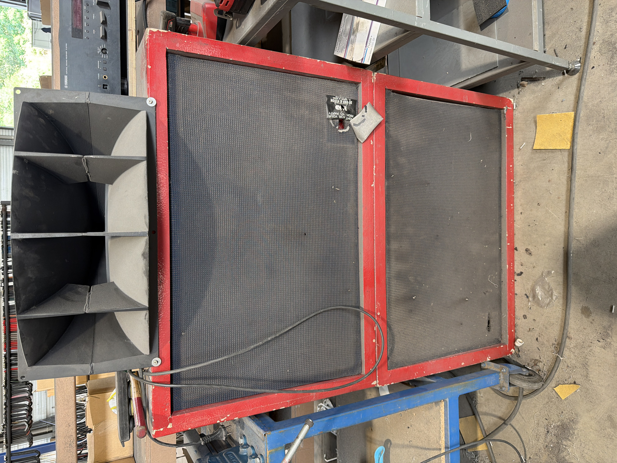 The image shows a large, multi-component speaker system in what appears to be a workshop or storage area. The speaker system consists of a large horn-loaded enclosure mounted on top of a two-tiered speaker cabinet. The cabinet has a red frame and black speaker grilles. There are various cables connected to the speakers and a Yamaha audio receiver is visible in the background. The environment is cluttered with tools, boxes, and other equipment. There is no indication of a specific location in the city of Mol, Belgium.