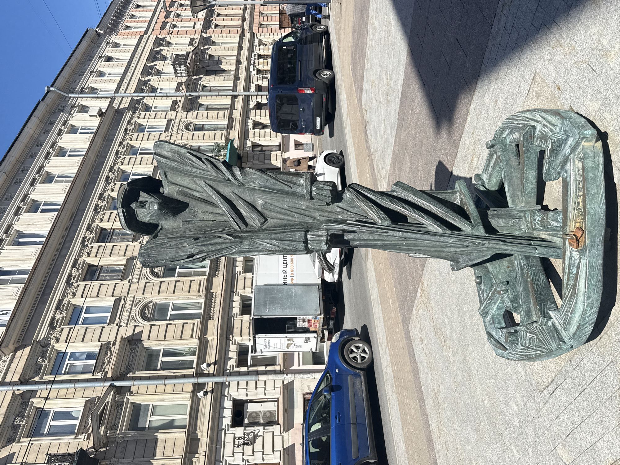 The image depicts a bronze sculpture of a figure standing on an ornate base on a sunlit street in Saint Petersburg, Russia. The sculpture, rendered in a somewhat abstract and angular style, features a tall, draped form with a visible head covered by a hat or hood. The base is circular and appears to be made of interconnected geometric elements.
In the background, a multi-story building with ornate architectural details, typical of Saint Petersburg's historical districts, lines the street. The building has numerous windows and decorative cornices.
Several vehicles are visible on the street. In the foreground, a blue car is parked. Behind it, a white delivery truck with its rear doors open is parked, displaying Russian text on its side. Further down the street, a dark blue minivan is in motion, and a white car is also visible. A few pedestrians are present, one walking away from the camera in the background, and another person standing near the minivan. The street is paved with a combination of asphalt and paving stones. Power lines and a lamppost are also visible. The overall impression is of an urban street scene on a clear, bright day.