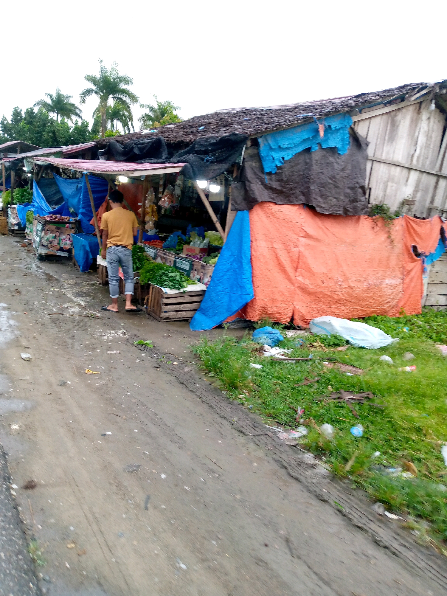 In Gampongpaya, Indonesia, an outdoor market stalls line a muddy dirt road. Several temporary structures with corrugated tin roofs and tarp coverings are visible, displaying various goods, likely produce, on wooden crates. A man wearing a yellow t-shirt and blue jeans stands facing away from the camera, looking at the market stalls. The ground is wet and appears to be a mixture of mud and gravel, with some scattered debris. Green vegetation and palm trees are visible in the background. The sky is overcast. The scene depicts a modest, functional marketplace in a rural or semi-rural setting.