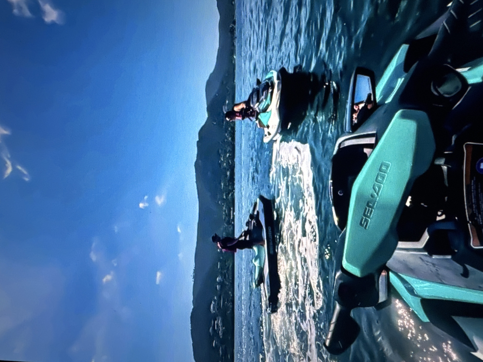 In a bright, sunlit scene, two individuals are riding jet skis on a body of water. The camera is positioned on a jet ski, looking forward, with the handlebars and front of the jet ski in the foreground. The handlebars are a teal color and have "SEA-DOO" branding visible. To the left, a person wearing a red life vest and a dark hat stands on a dark-colored jet ski, creating a spray of white water. To the right, a person wearing a red life vest sits on a teal and black jet ski. In the background, a lush, green hillside covered in buildings stretches across the horizon under a clear blue sky with scattered white clouds. The water is choppy, with white foam and ripples generated by the jet skis. The scene appears to be a recreational activity taking place on a sunny day.