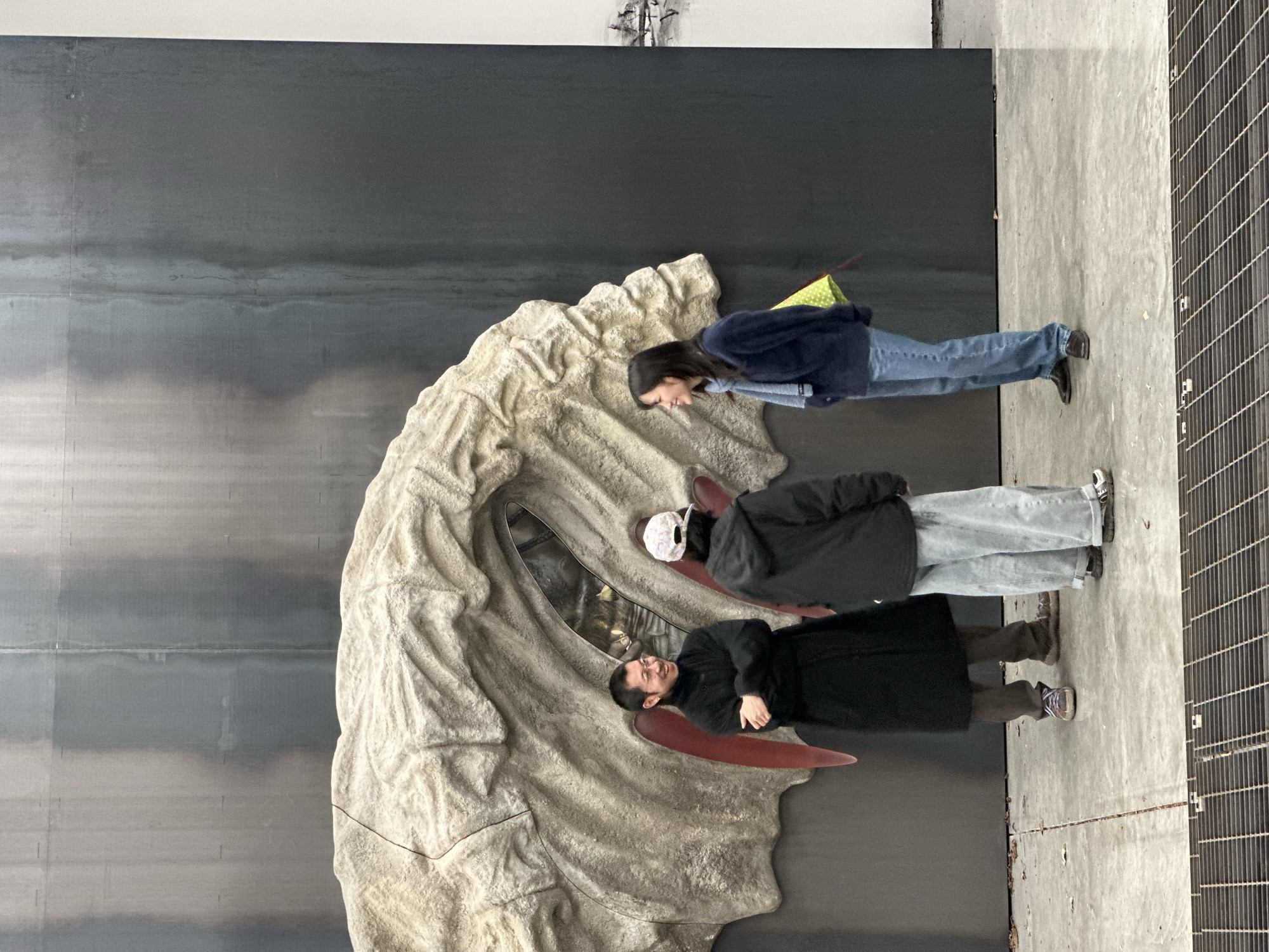 The image displays three individuals within an indoor contemporary art exhibition space in London, United Kingdom. A large, light-grey, textured sculpture, resembling an organic form or stylized rock formation, occupies the center-left. This sculpture is mounted against a dark grey, vertically paneled wall, and features a central recessed opening with visible reflective elements, flanked by two downward-pointing red, conical protrusions.
From left to right:
1. A male individual, wearing glasses, a black high-necked tunic, brown trousers, and sneakers, stands with arms crossed, smiling and looking towards the right.
2. An individual, mostly seen from the back, wears a dark hooded jacket, light-wash wide-leg jeans, and a white patterned baseball cap.
3. A female individual, wearing a dark blue sweater, blue jeans, a light blue scarf, and dark shoes, stands in profile, smiling towards the left. She carries a yellow-green polka-dotted bag over her left shoulder.
The individuals appear engaged in conversation. The floor is light grey concrete with scattered debris. A metal grate structure is visible in the immediate foreground. The background wall exhibits vertical lines and tonal variations consistent with large panels.