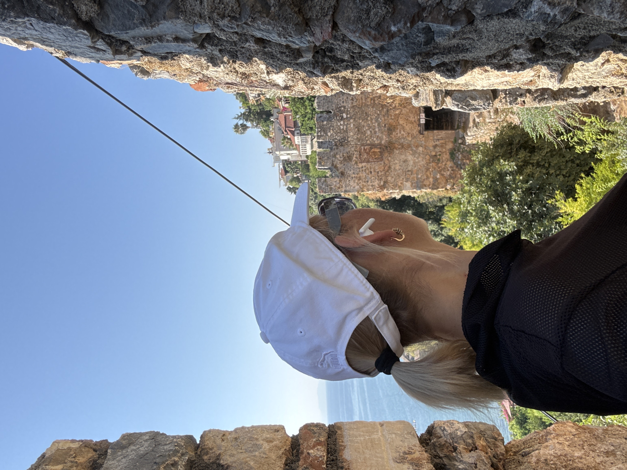 A woman wearing a white baseball cap and sunglasses is viewed from behind, looking out over a city landscape from a stone structure. She has blonde hair pulled back in a ponytail and is wearing a black mesh shirt. White earbuds are visible in her ears, and a gold hoop earring is on her right ear. The background reveals a body of water, a stone fortress, and buildings on a hill, consistent with a view from Alanya Castle in Alanya, Turkey. The sky is clear and blue. The image is framed by the rough texture of stone walls on either side, suggesting the woman is positioned within a window or opening of the castle.