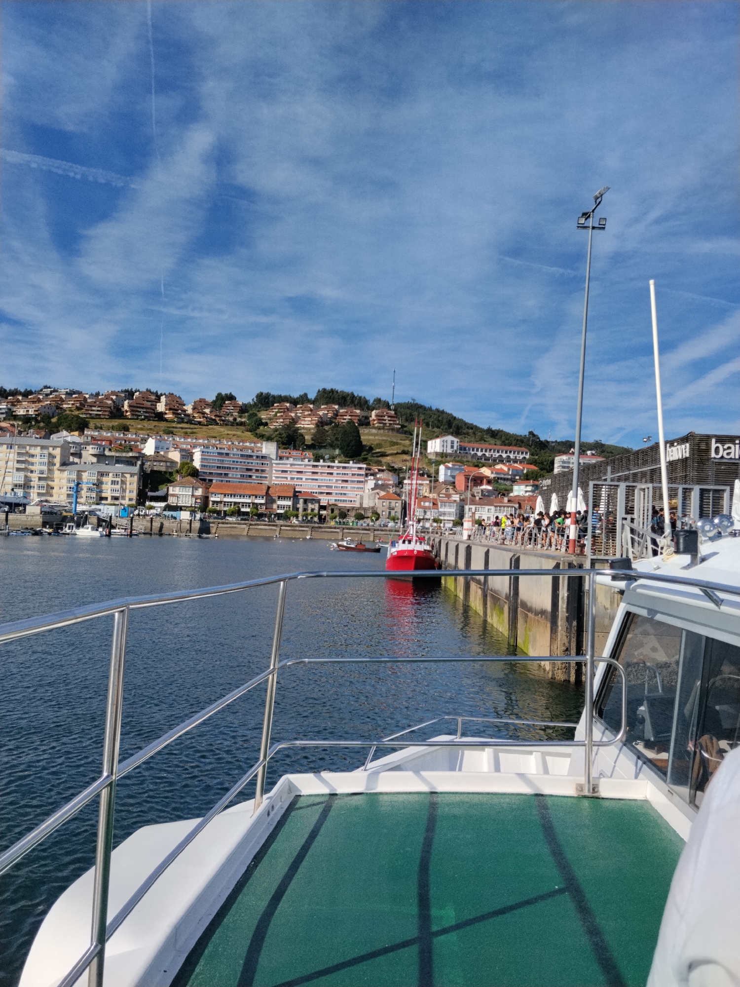 The image shows a view from a boat in the harbor of Baiona, Spain. In the foreground, a portion of the boat's deck and railings are visible. The water in the harbor is dark blue and slightly rippled. Across the water, the town of Baiona is built on a hillside, with many buildings and houses. A red and white boat is docked in the harbor, and a pier with people walking on it is also visible. Several other boats are moored in the harbor, and a large building with the word "bai" on it is situated near the pier. The sky is blue with scattered white clouds.