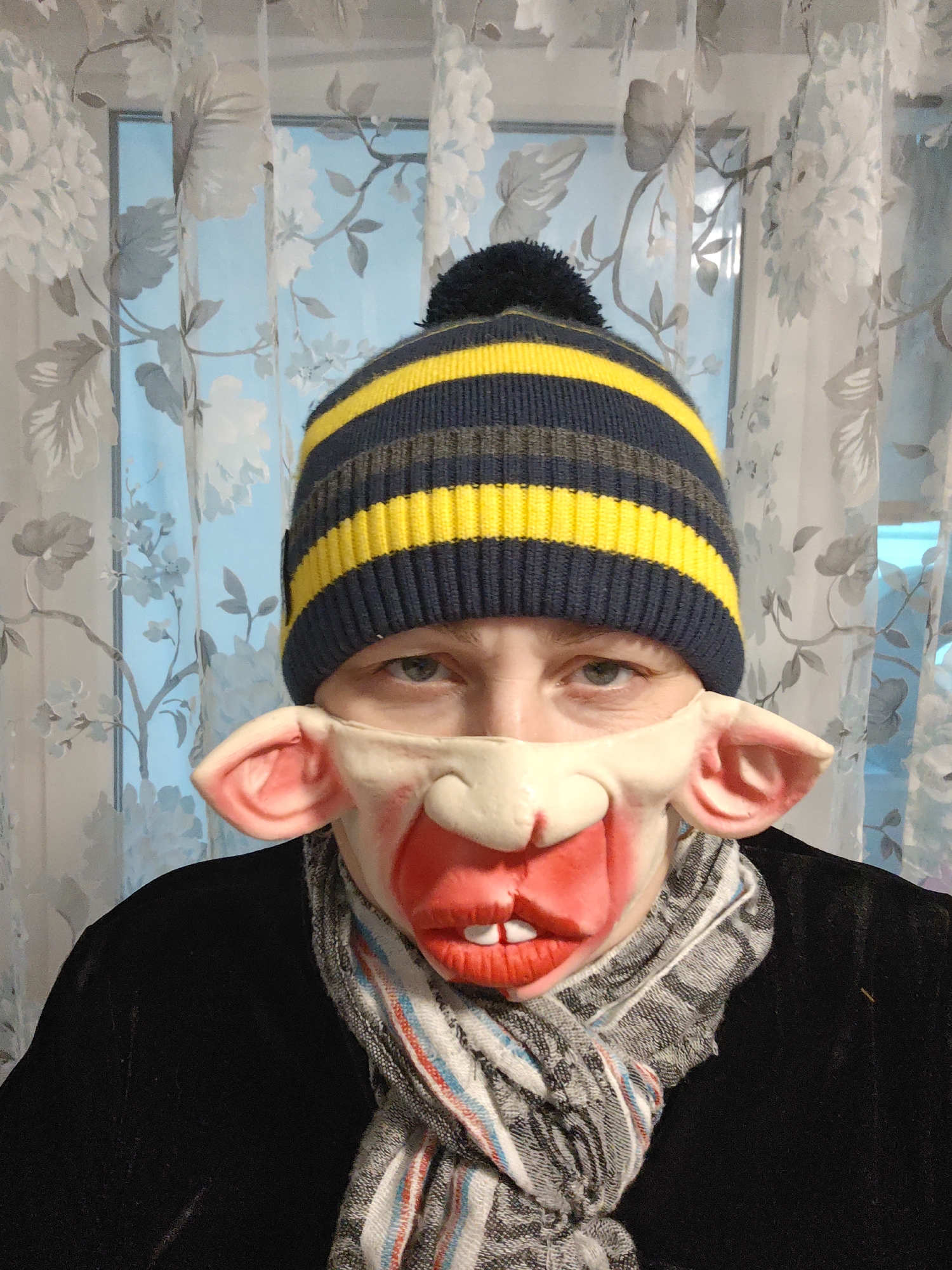 A person is wearing a blue and yellow striped knit cap and a pig mask. The mask covers the lower half of their face and has large, pink ears. A gray and white scarf is wrapped around their neck. They are wearing a black jacket or coat. The background consists of a window covered with a floral patterned curtain. The location is implied to be Markovskii, Russia, but this detail is not discernible from the image itself.