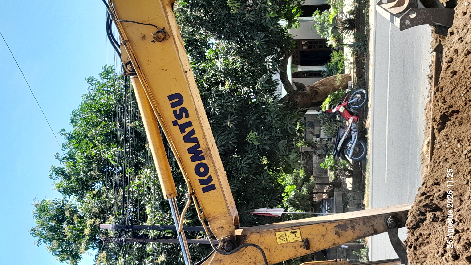 The image depicts an outdoor scene on a sunny day. A yellow excavator boom, prominently displaying the blue "KOMATSU" logo, is in the foreground, angled from the lower left to the upper right. To the left of the excavator arm, there's a warning sign with a triangle and an exclamation mark, and below it, symbols indicating movement and danger. A red and silver motorcycle is parked on the side of a road in the middle ground, with lush green trees and foliage in the background. A white building with columns and a dark-colored door is partially visible behind the trees. A small Indonesian flag is planted in the ground near the motorcycle. The ground in the immediate foreground is composed of dark brown soil, and a timestamp in the lower left corner reads "25 Agustus 2026 11.25". The scene is suggestive of construction work or maintenance, potentially in a residential or semi-rural area of Jomblang, Indonesia.
