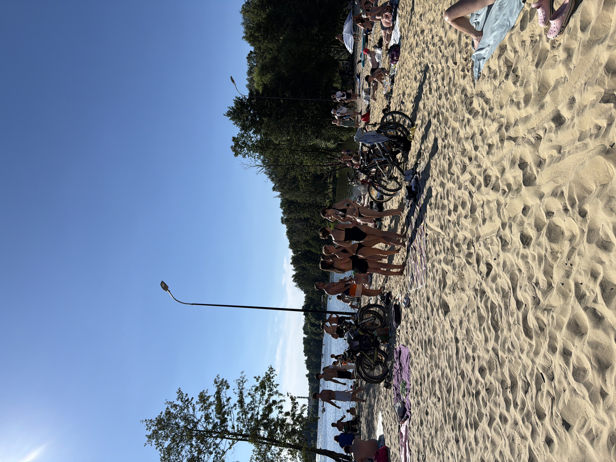 This image depicts a crowded beach on a sunny day in Malakhovka, Russia. The beach is filled with people, many of whom are sunbathing, swimming, or standing and conversing. Several bicycles are parked on the sand, suggesting that beachgoers arrived by bike. The environment includes a body of water in the background, with trees lining the shore. The sky is clear blue. There are beach towels and bags scattered across the sand, indicating a leisurely atmosphere.
