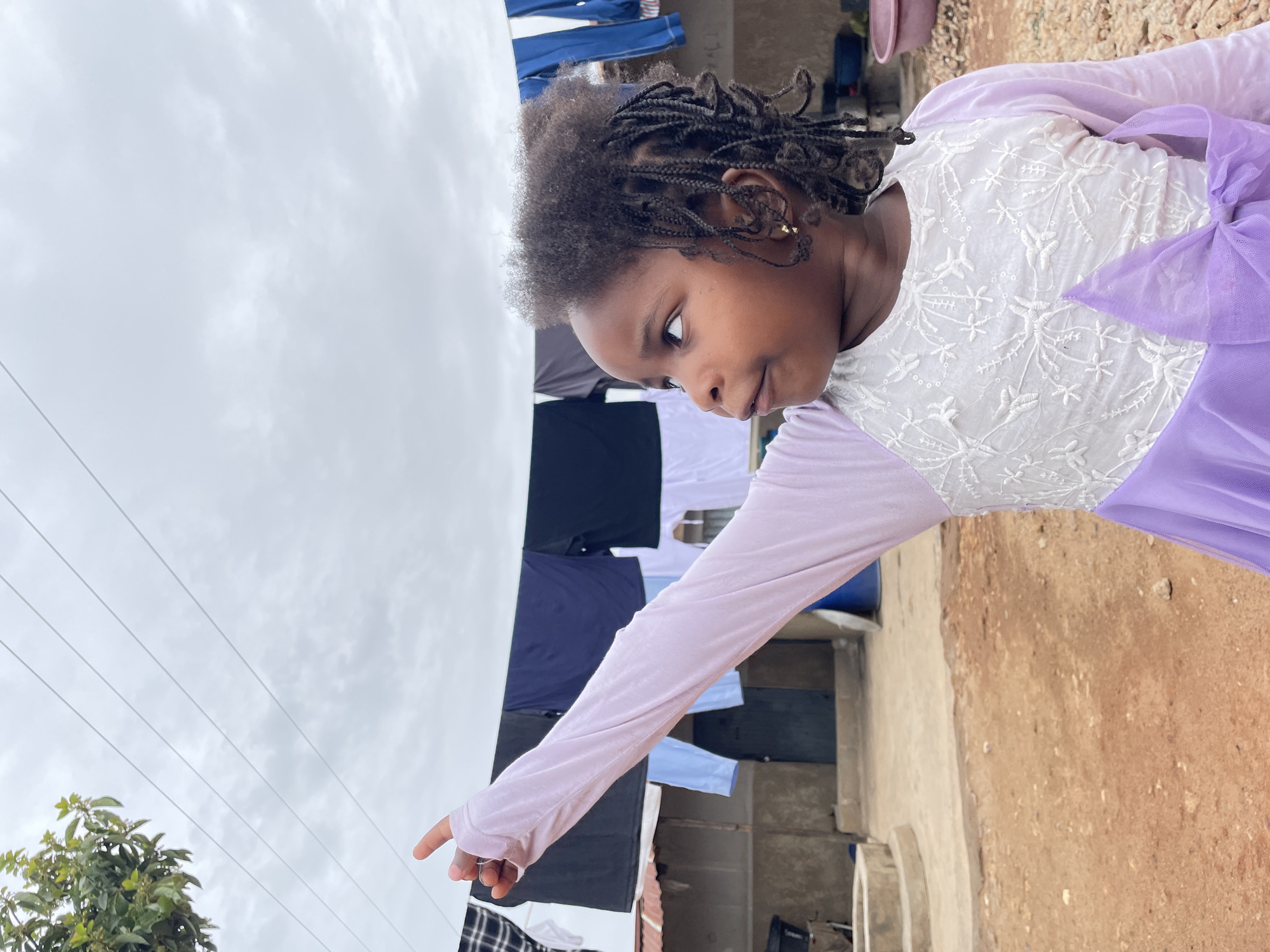 A young girl stands outdoors, seemingly pointing towards the sky. She has dark hair styled in braids and is wearing a dress with a white embroidered bodice and a light purple skirt and long sleeves. Behind her, laundry hangs on a clothesline. The ground is dirt. In the background, concrete structures are visible. Based on the context, the location is likely Jos, Nigeria.
