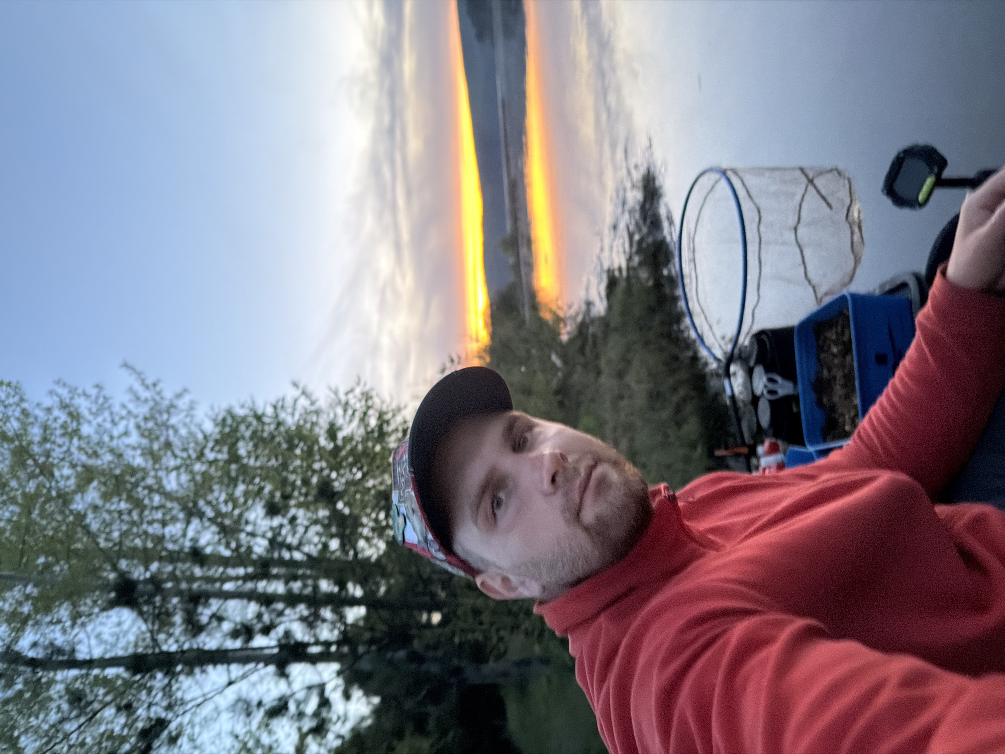 A selfie of a man in a red zip-up shirt and a baseball cap, taken outdoors. The background features a body of water reflecting a sunset sky, with green trees and foliage visible on the shoreline. To the right of the man are fishing accessories, including a net and a blue container.
Although not definitively confirmed, based on the sunset over water and the presence of fishing equipment, it is likely that the photo was taken near the Kama River, which runs through Chaykovsky, Russia.